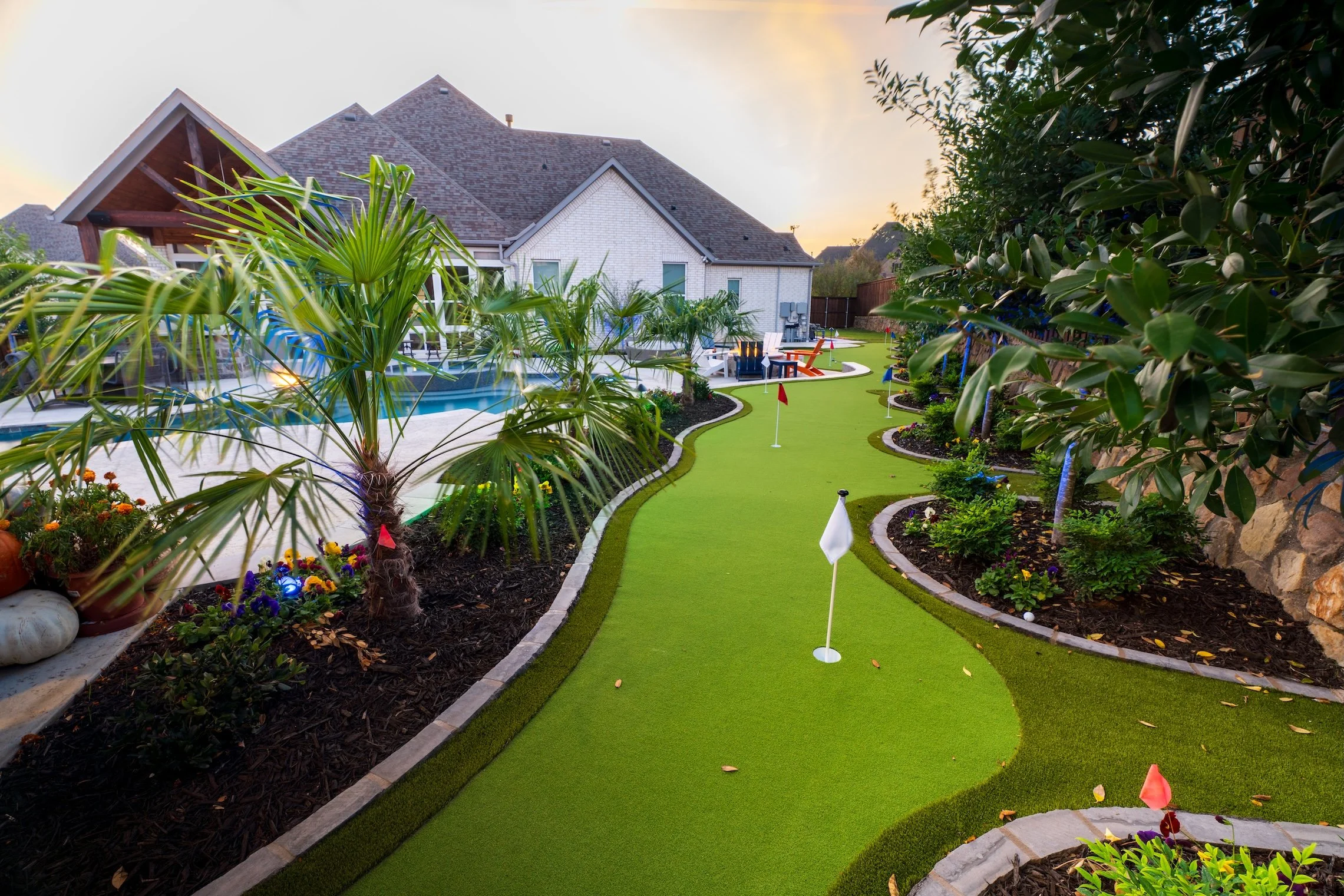 Backyard mini golf course with green turf, flags, plants, flowers, and a pool in the background at sunset.
