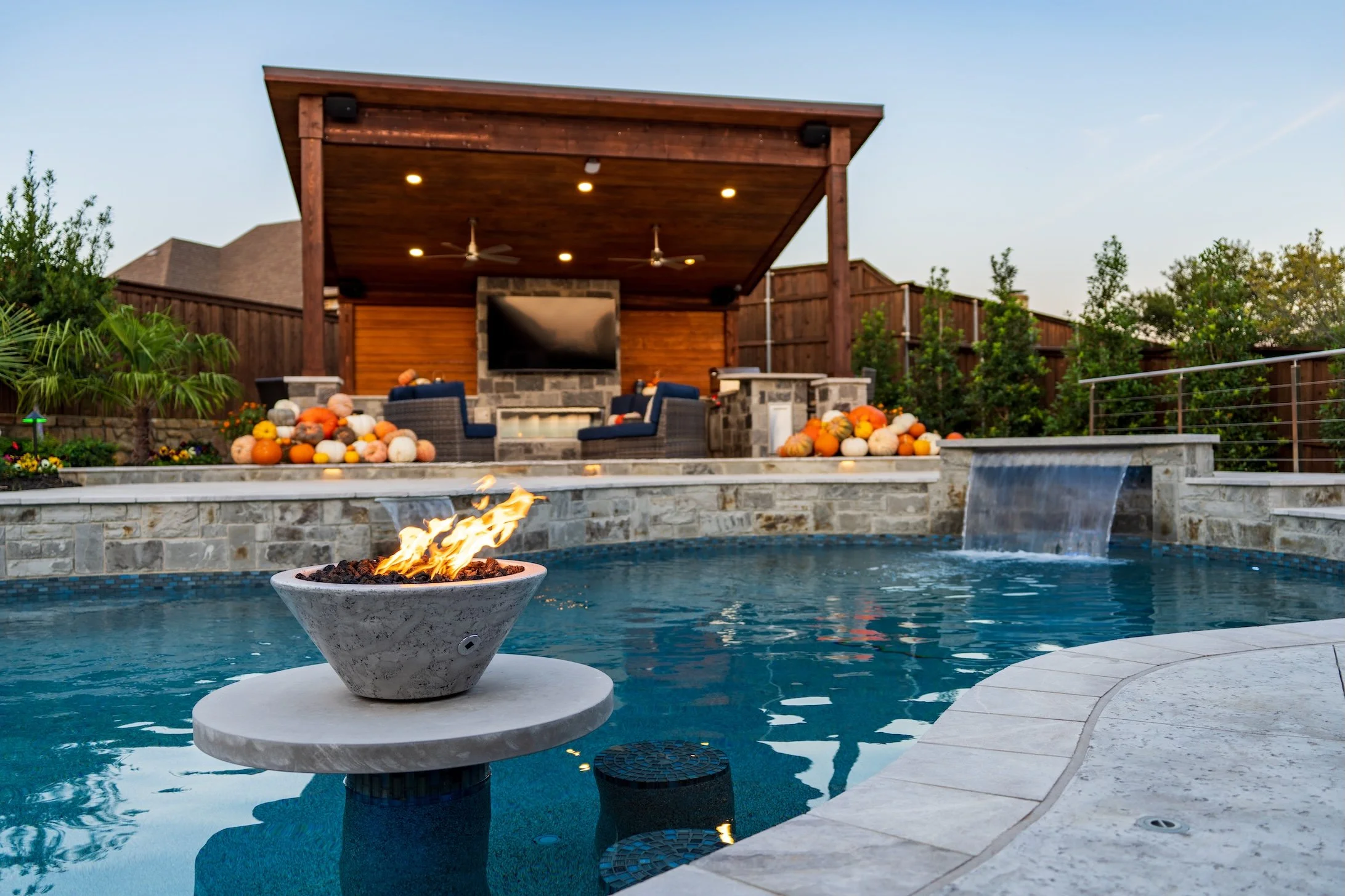 Backyard pool area with a stone fire feature, surrounded by fall pumpkins and gourds, with a covered outdoor living space with a TV, seating, and a waterfall feature.