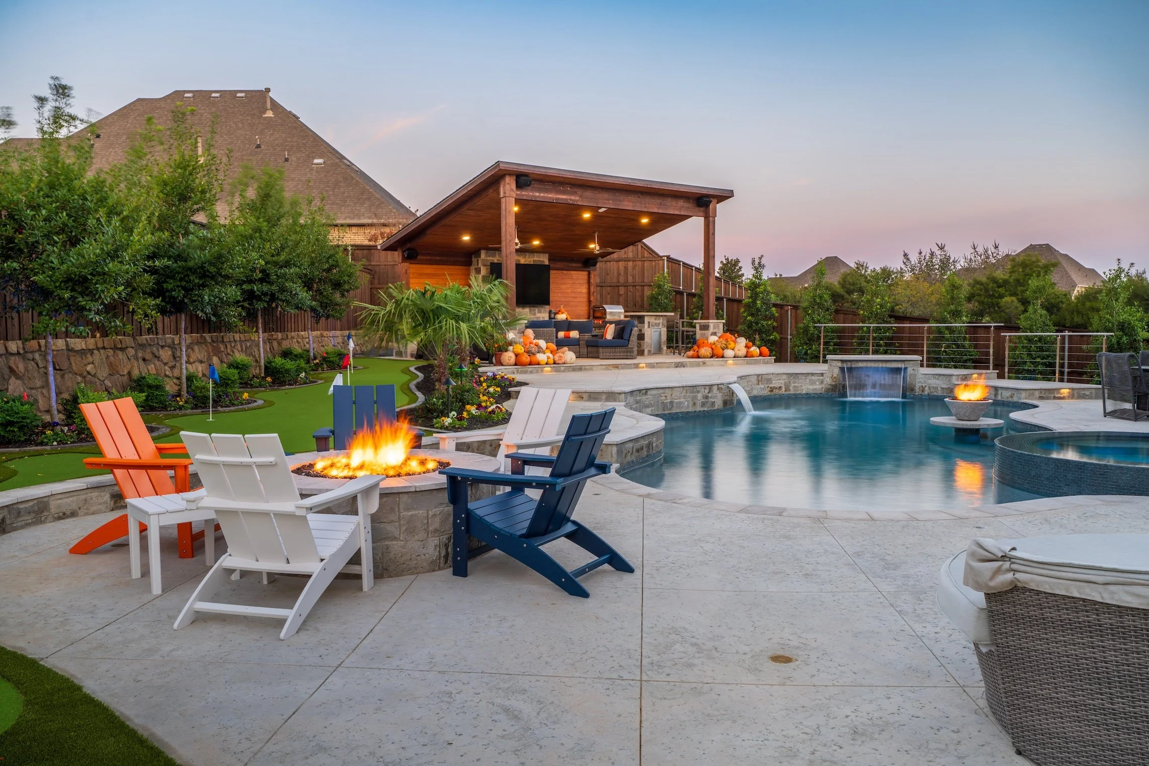 A backyard with a swimming pool, fire pit with chairs, a covered outdoor seating area decorated with pumpkins, trees, and greenery during dusk.