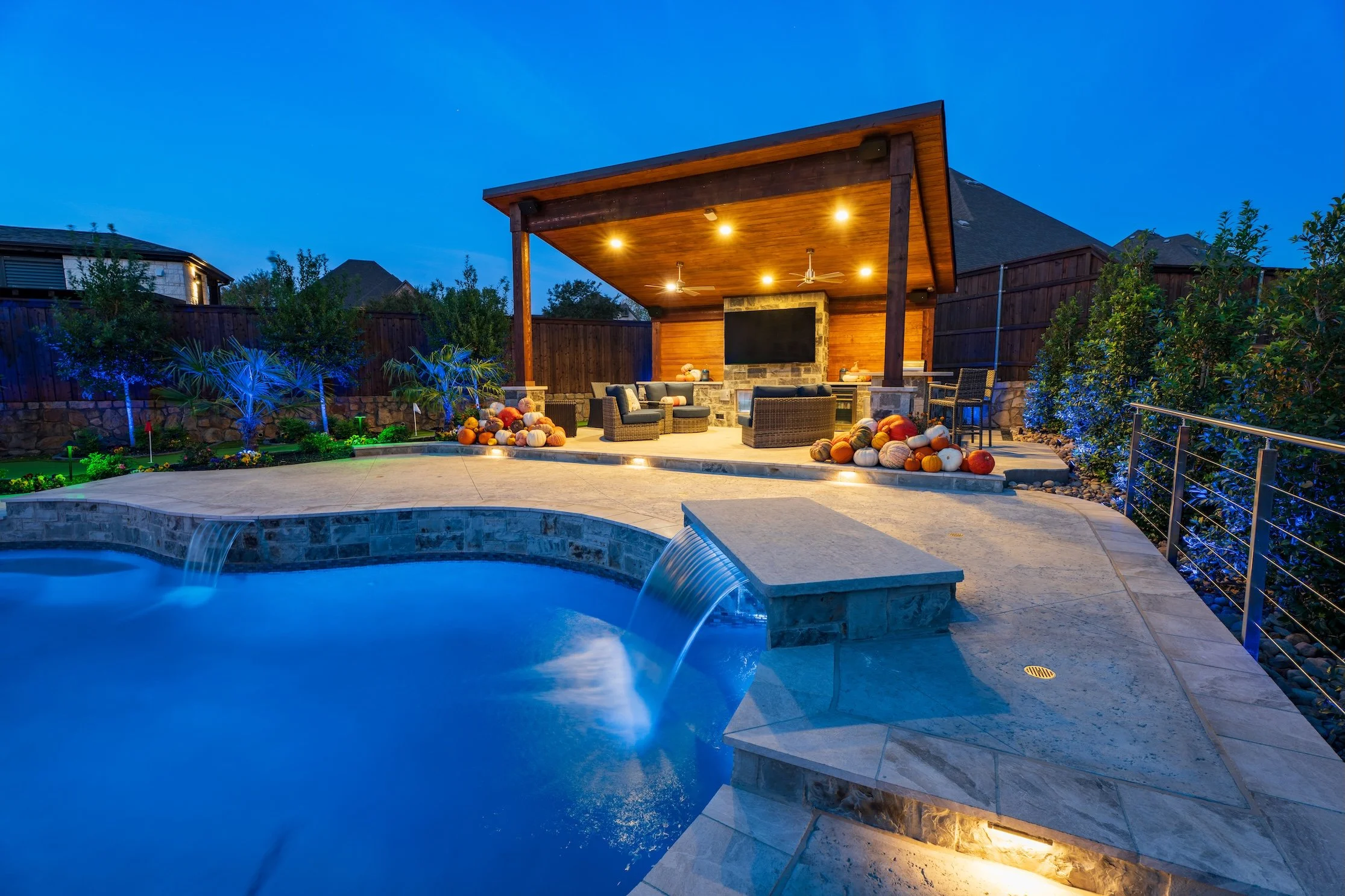 Backyard with a swimming pool, outdoor covered patio with seating, fireplace, and pumpkins for decoration, illuminated during evening.