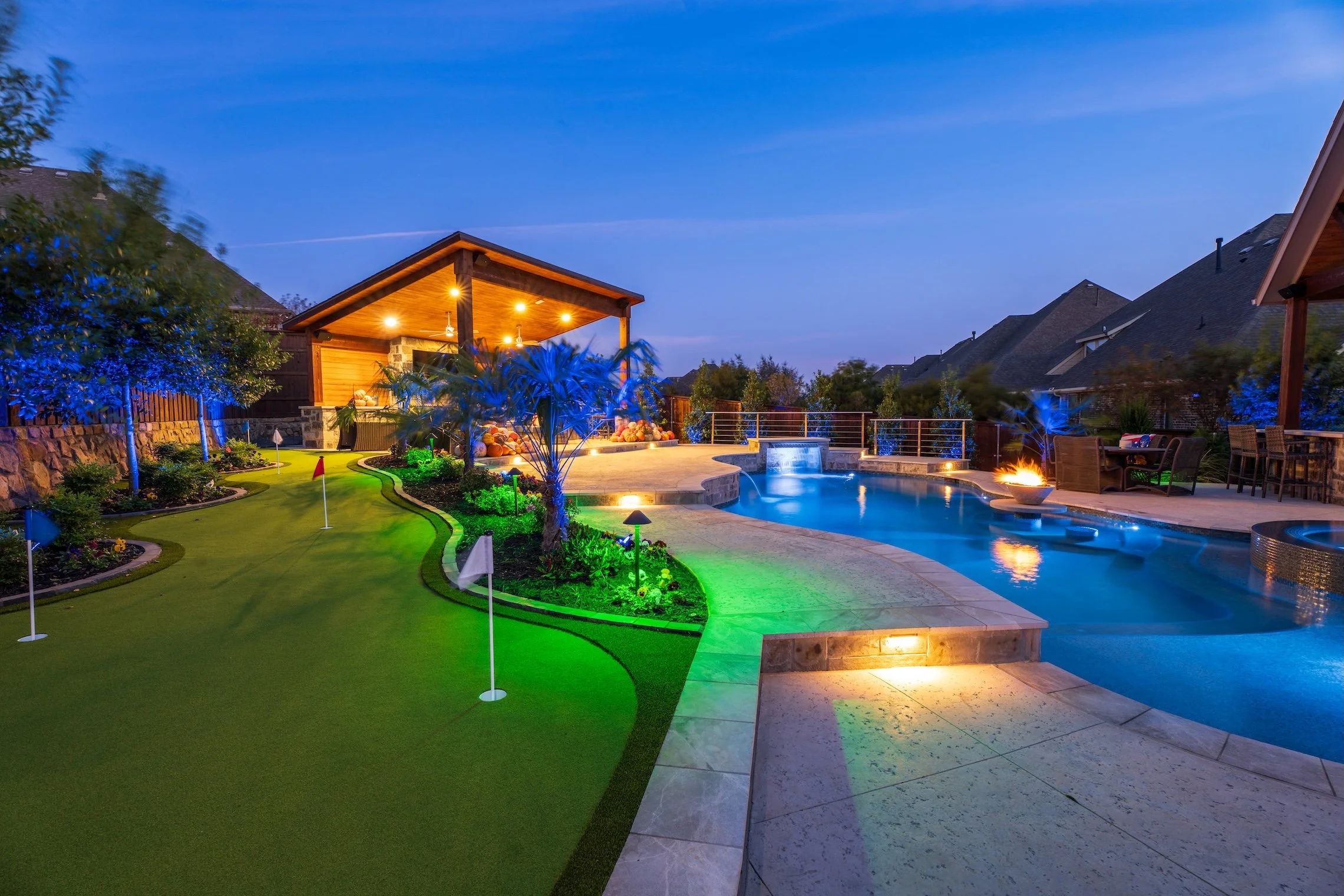 Nighttime view of a backyard with a lit swimming pool, fire feature, outdoor seating area, and a putting green with flags, surrounded by landscaped plants and trees.