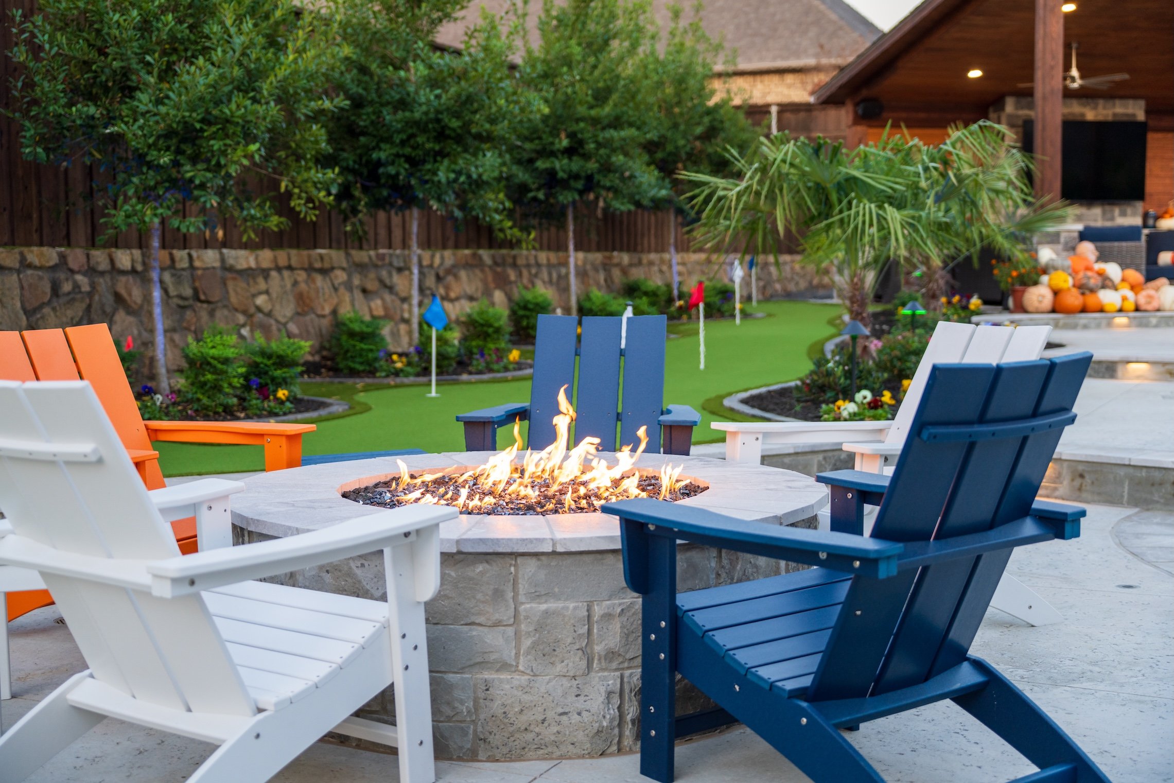 Backyard patio with a fire pit surrounded by four colorful Adirondack chairs in white, orange, dark blue, and light blue. In the background, there is a lush green lawn with small trees, plants, and decorative flags, bordered by a stone wall. To the r