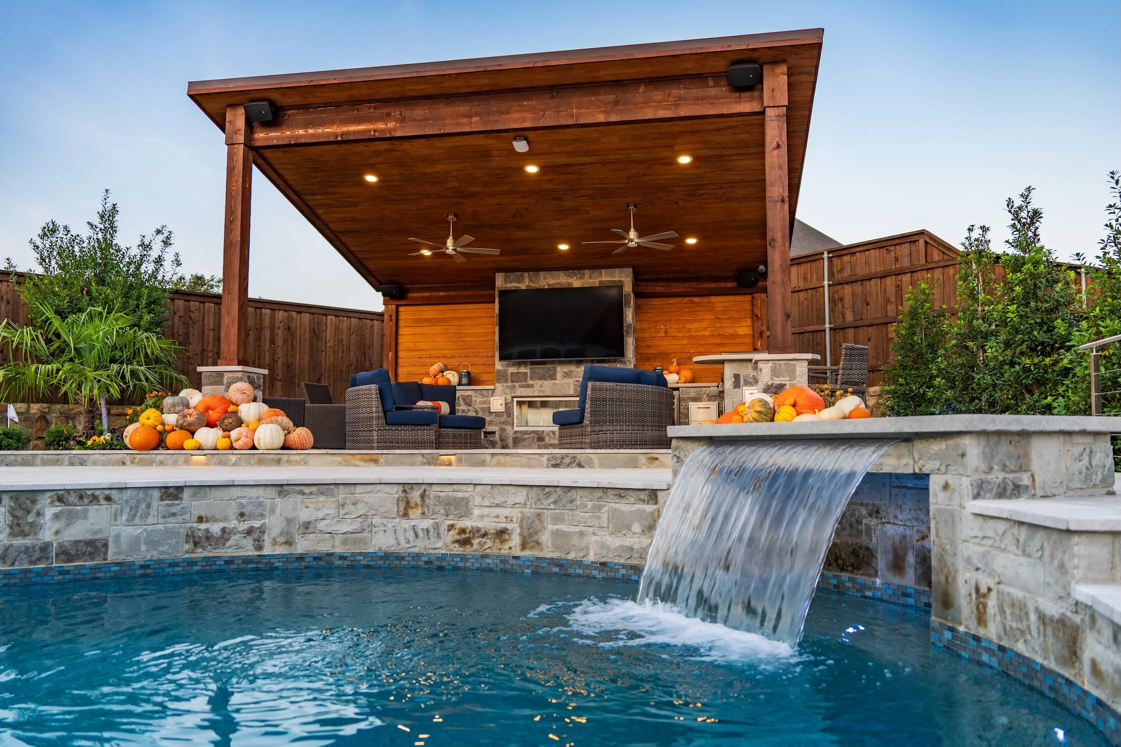 Backyard pool area with a waterfall, outdoor seating, a mounted flat-screen TV, and fall pumpkins and gourds for decoration.