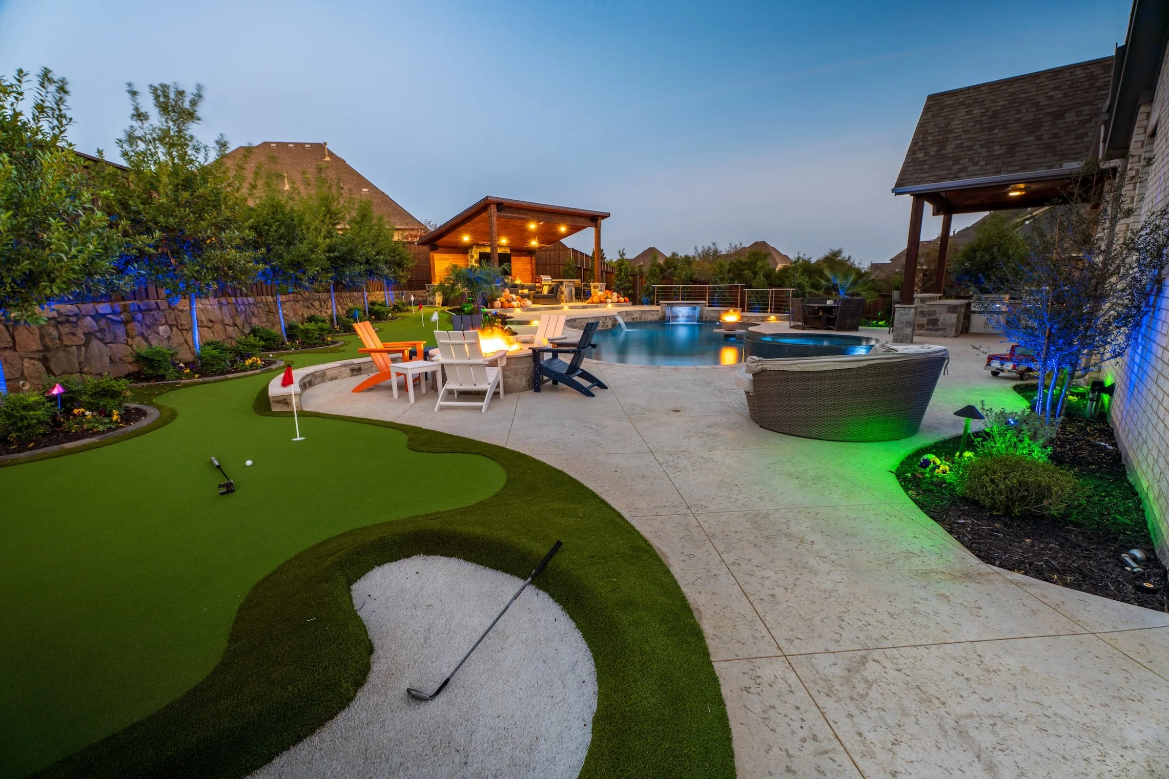 Backyard with a mini golf putting green, swimming pool, and a covered outdoor seating area at dusk, illuminated with colorful lights.