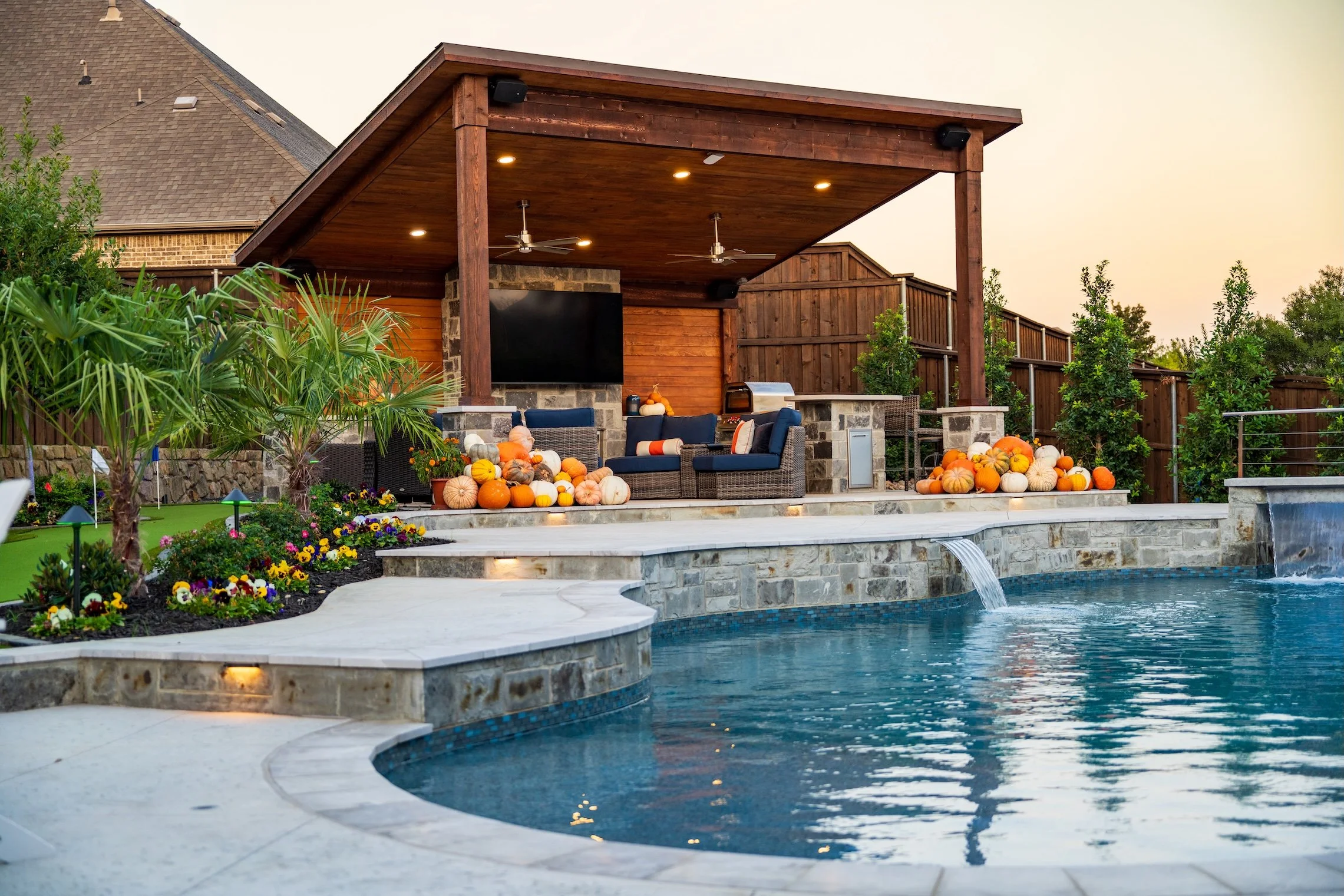 Backyard with a swimming pool, a covered patio area with outdoor furniture, pumpkins for fall decor, and landscaped garden.