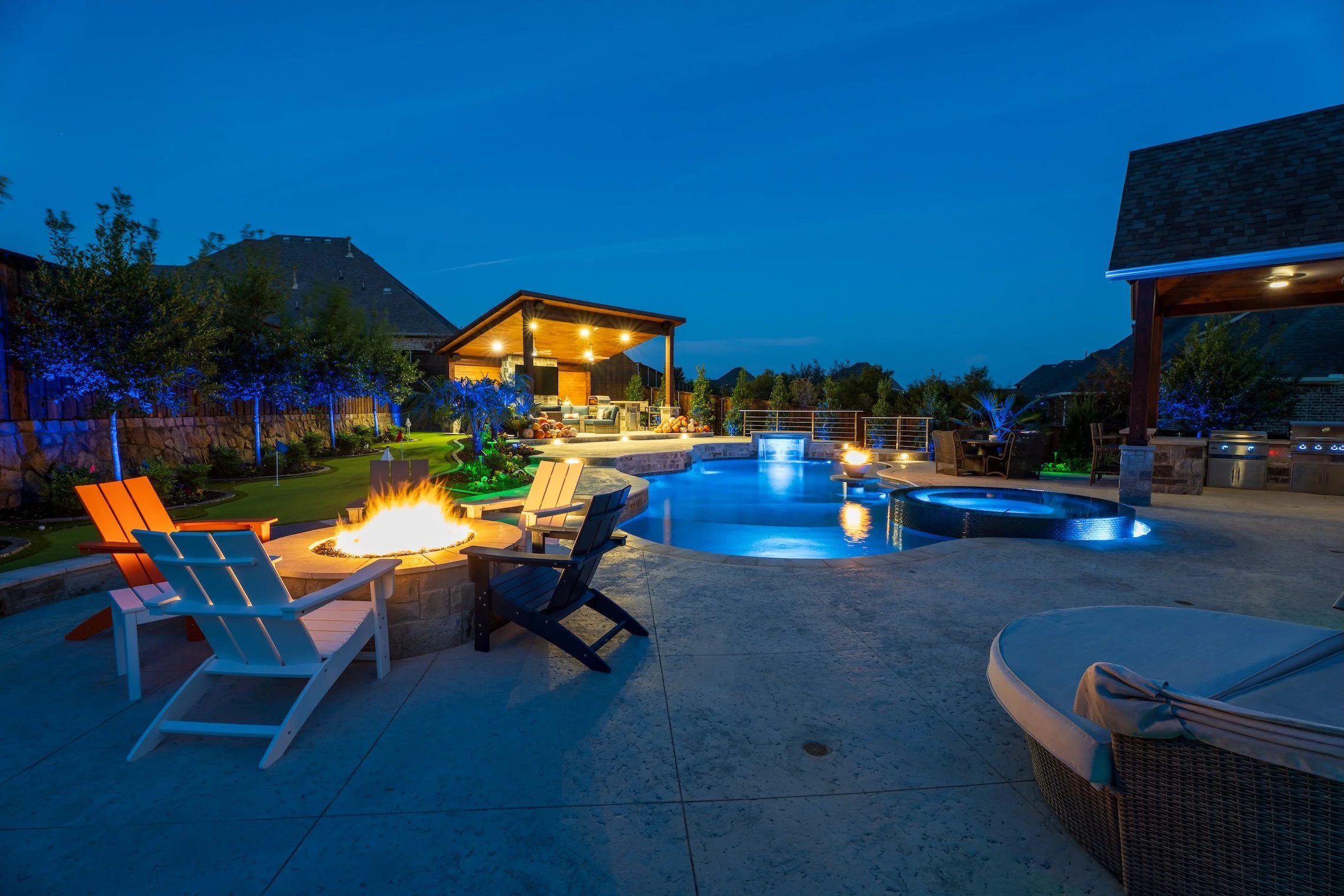 A nighttime view of a luxurious backyard with a swimming pool and hot tub, illuminated by fire pits. There are several colorful chairs around a fire pit in the foreground and a covered lounge area in the background with seating and outdoor kitchen am
