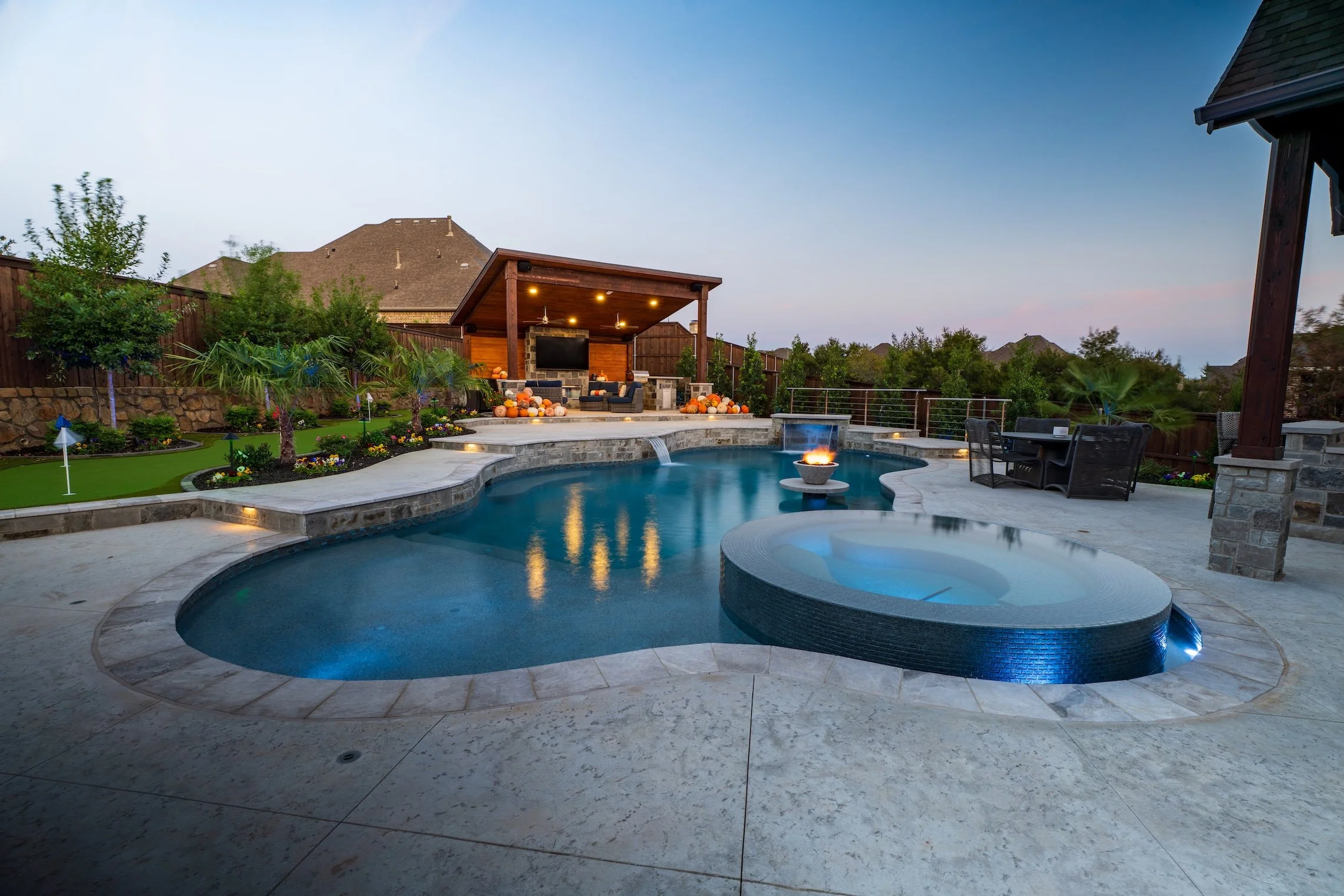 Backyard with swimming pool, hot tub, fire pit, outdoor seating, and a covered patio area with TV, surrounded by trees and a wooden fence at dusk.
