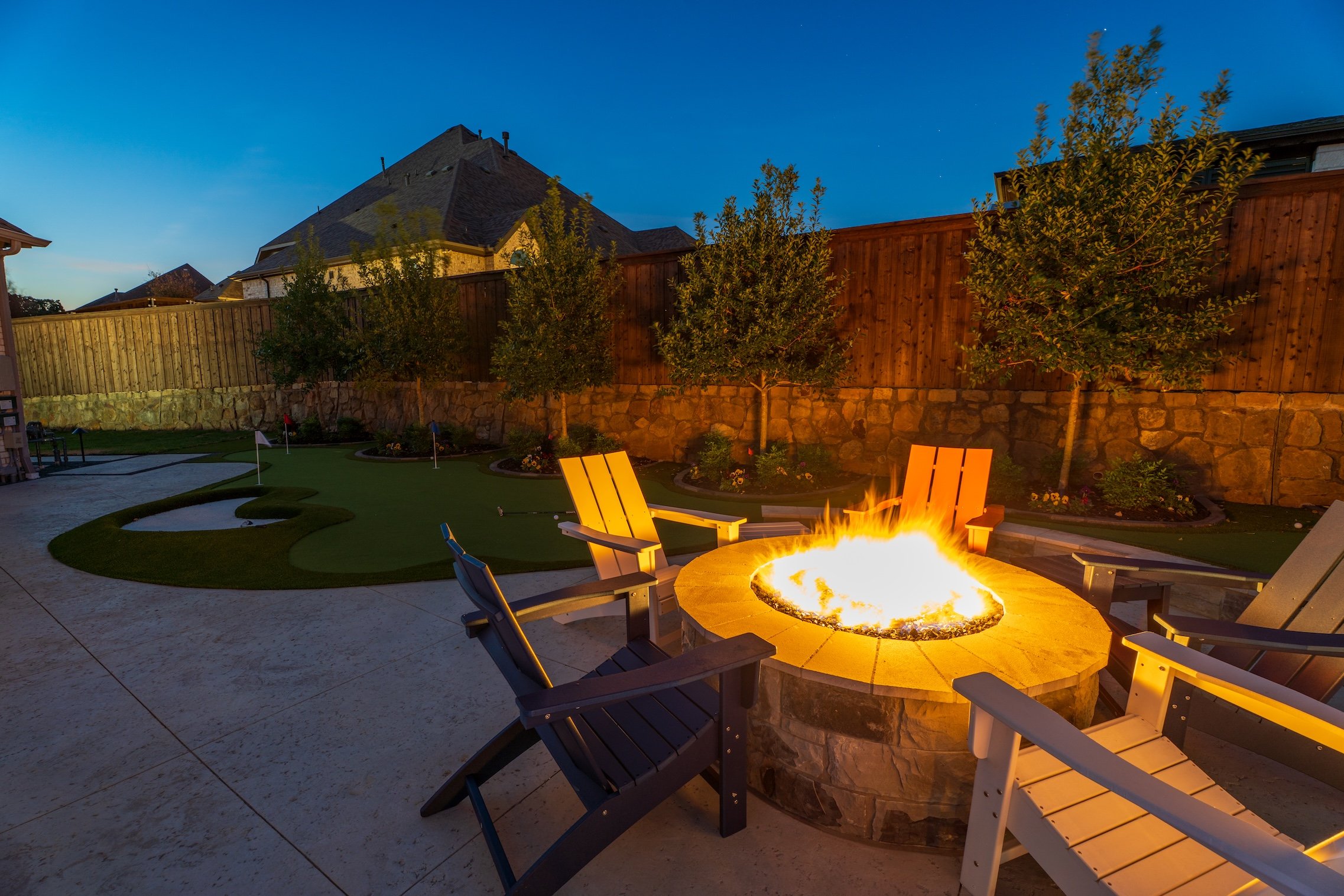 A backyard at dusk with four chairs surrounding a lit fire pit, small trees, a miniature golf course, a stone fence, and a clear evening sky.