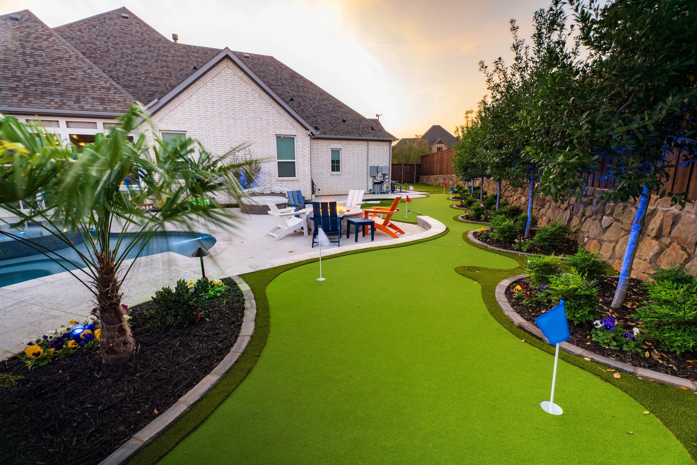 A backyard with a putting green, picnic table, Adirondack chairs, small pool, and a landscaped yard with trees and flowers at sunset.