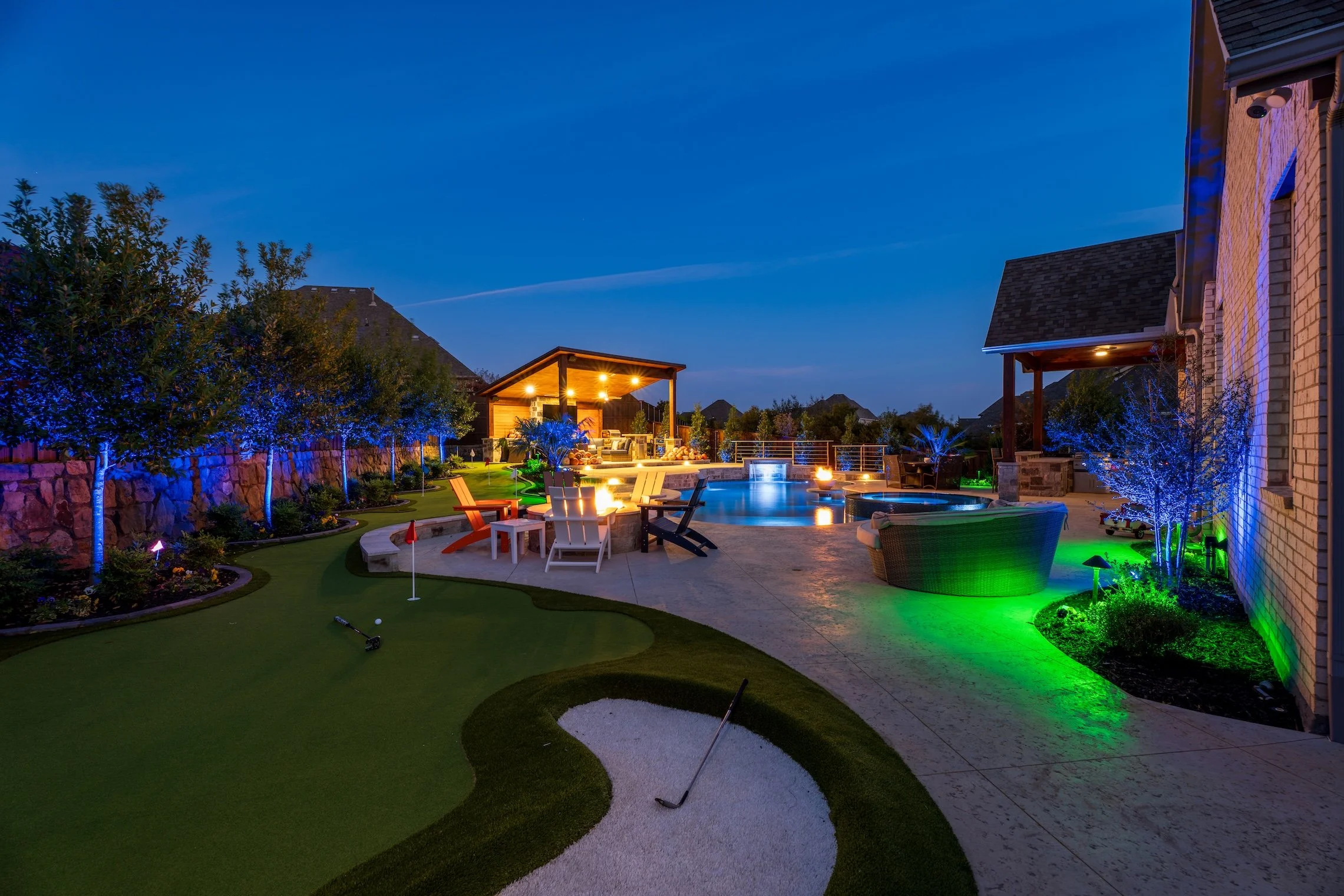 Nighttime view of a backyard patio with a swimming pool, outdoor fireplace, lounge chairs, and trees illuminated with colorful lights.