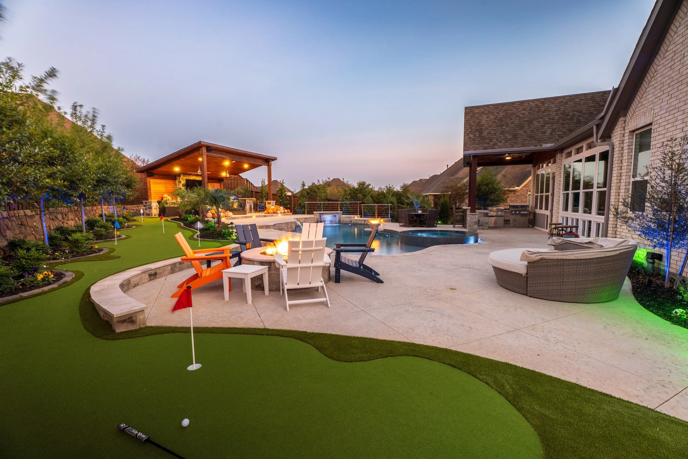 Backyard with putting green, fire pit, pool, hot tub, seating area under covered patio, and outdoor kitchen with counter and grill.