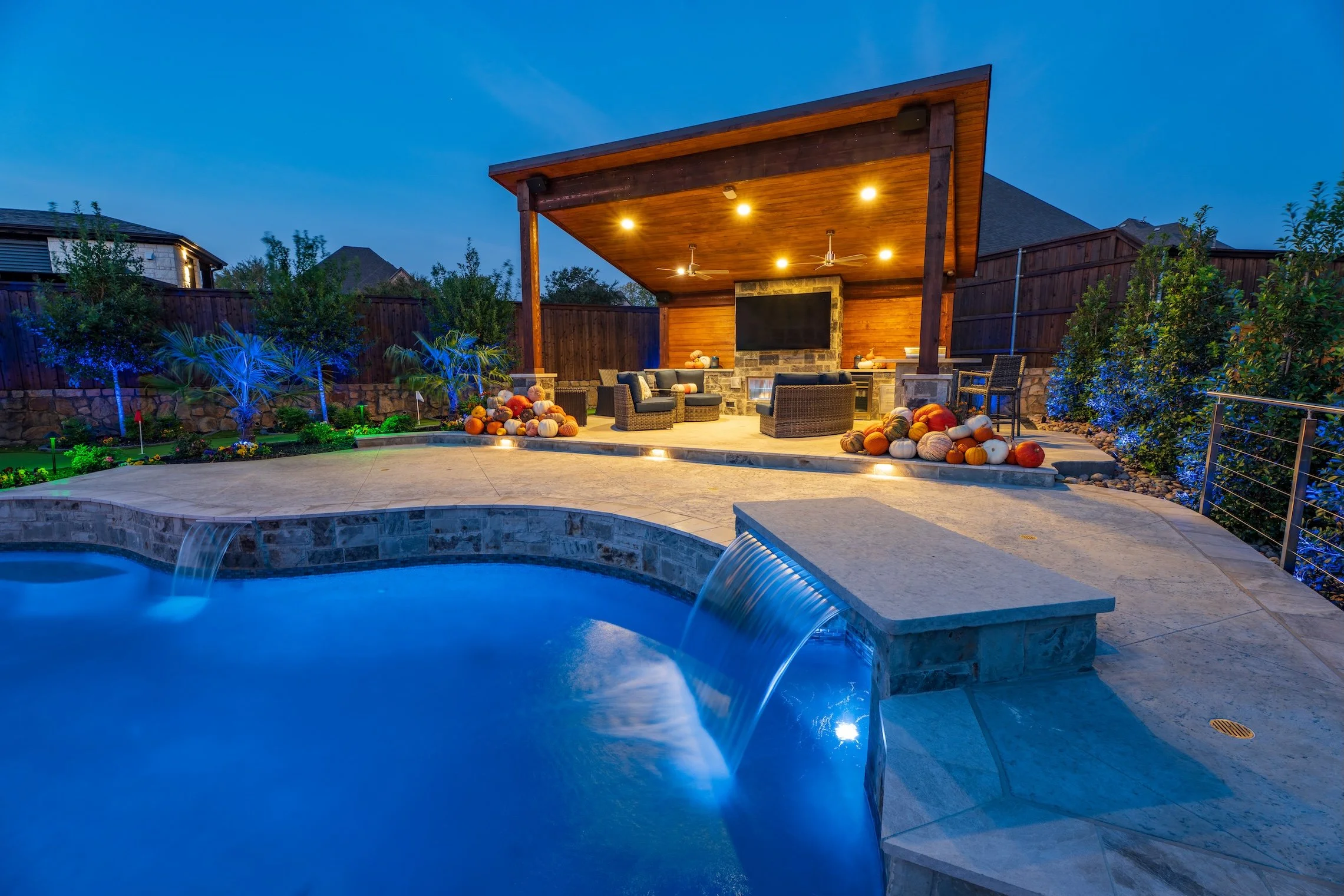 A backyard with a swimming pool and a covered outdoor seating area at dusk. The seating area has a stone fireplace and a TV, decorated with pumpkins. The pool has waterfalls, and the yard is landscaped with plants and trees.