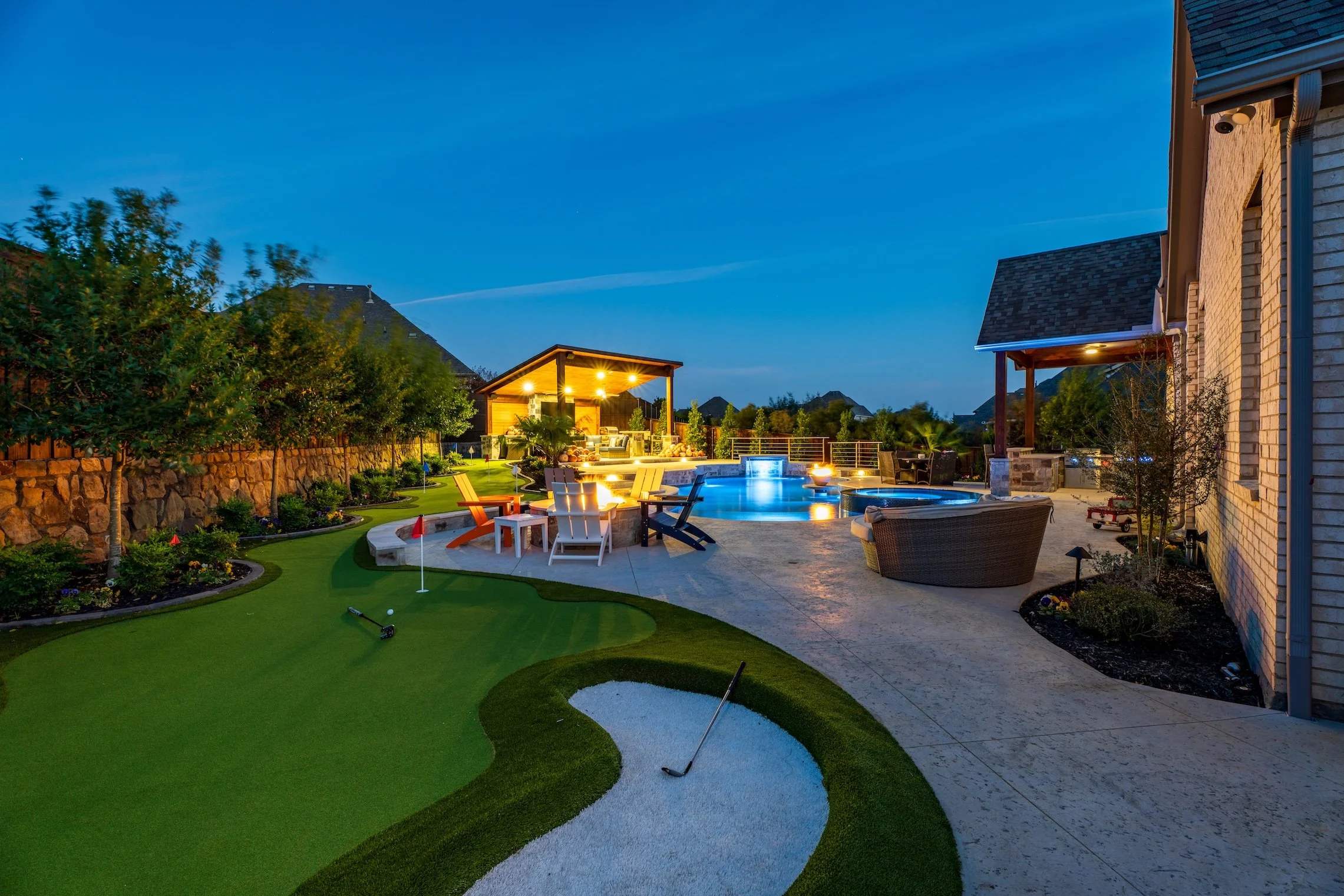 A backyard at dusk featuring a putting green, a sand bunker, a patio with outdoor furniture, a swimming pool with lighting, a hot tub, and a covered outdoor living area with string lights, surrounded by trees and landscaping.