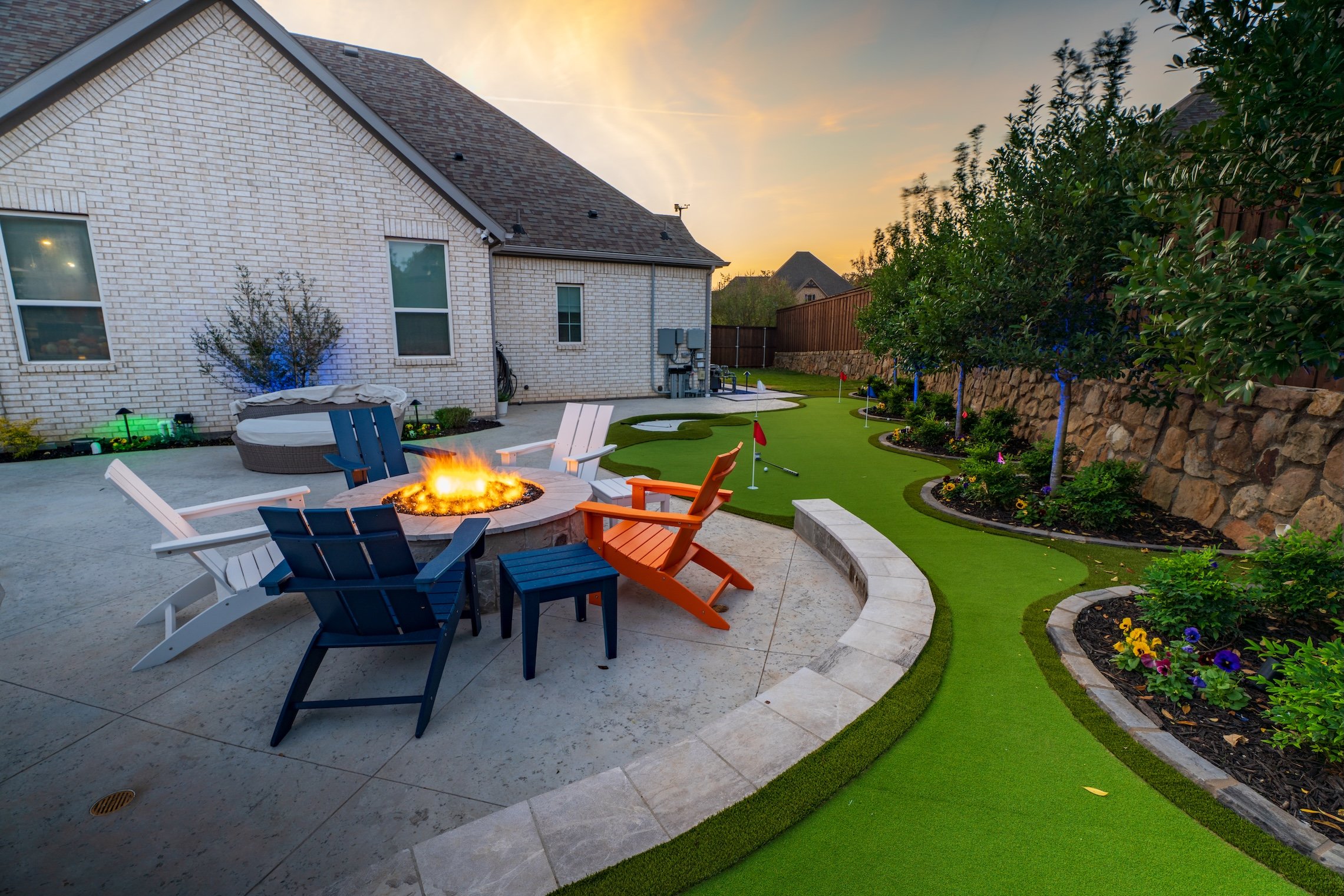Backyard with patio seating around a fire pit, putting green, trees, and flower beds at sunset.