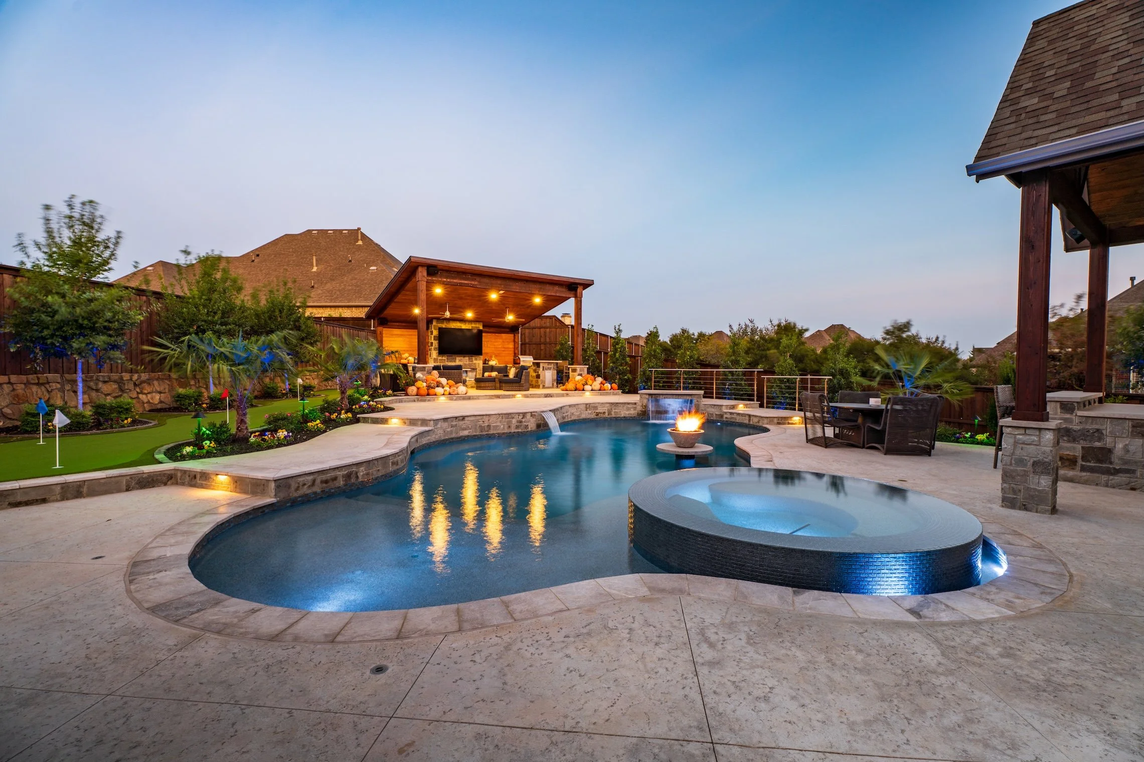 Backyard with a swimming pool, hot tub, outdoor seating, fire feature, and a covered patio area during dusk.