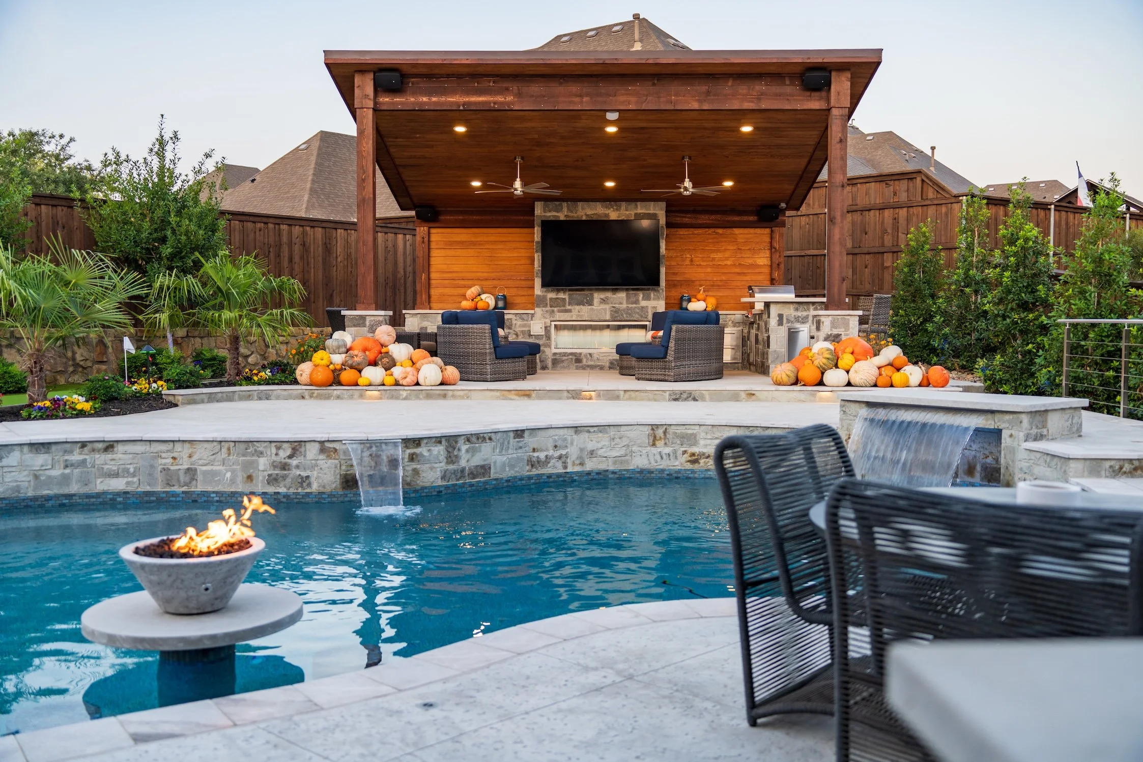 A backyard with a swimming pool, a fire pit, and a covered outdoor living area with a flat-screen TV. There are pumpkins and autumn decorations around for fall.