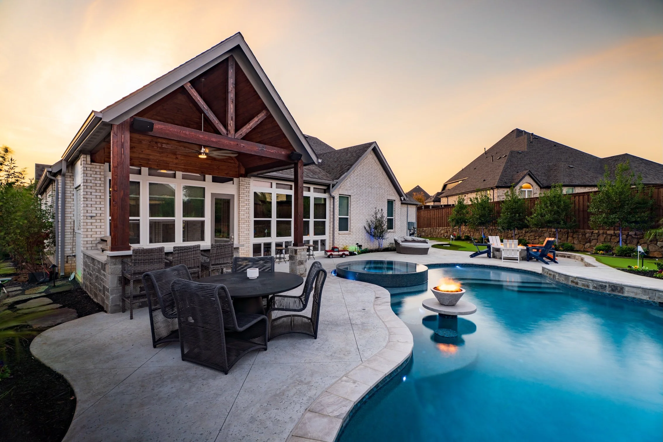 A backyard with a swimming pool, fire feature, patio furniture, and a house with large windows at sunset.