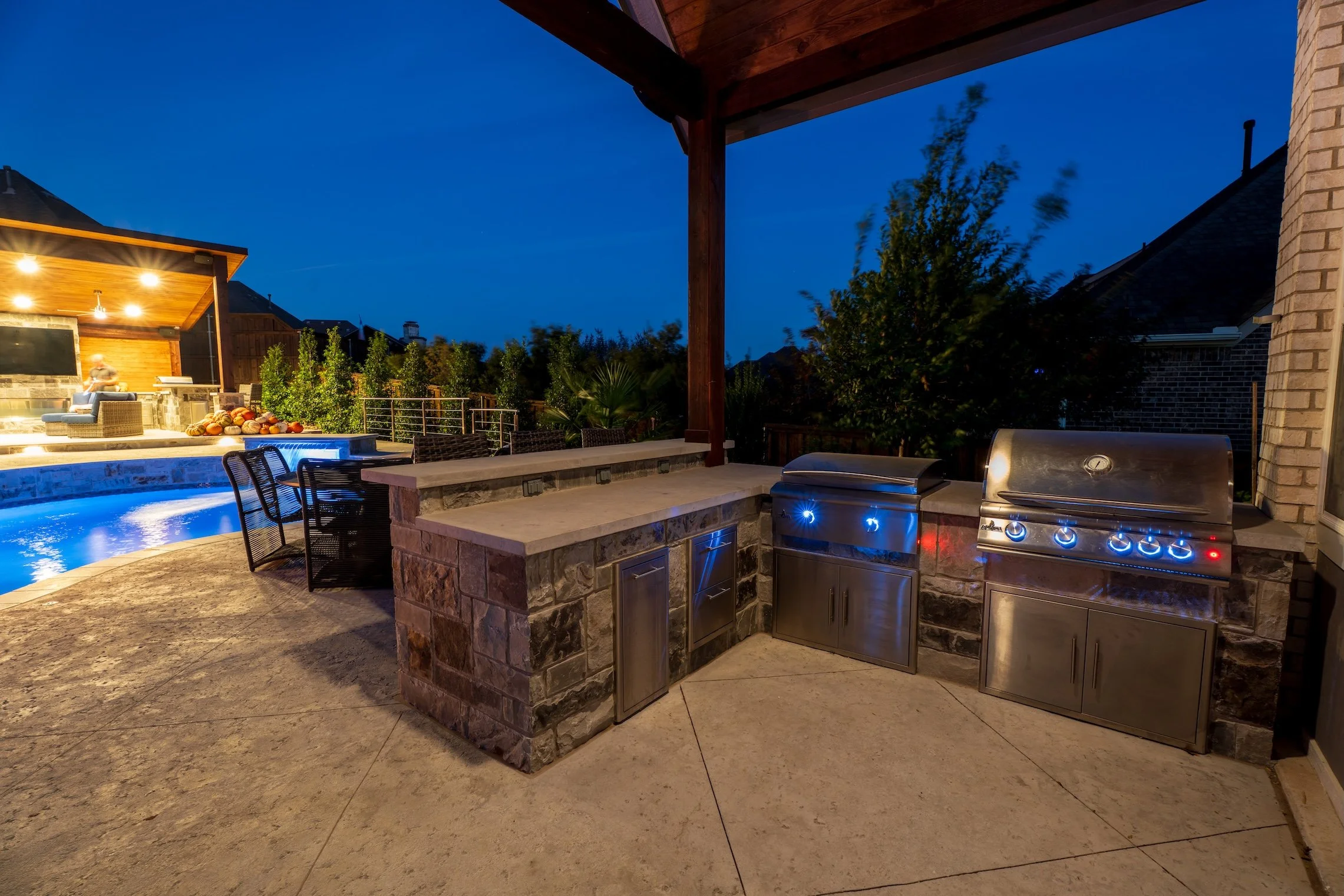 Outdoor kitchen area with stainless steel grills, stone countertops, and a view of a backyard pool with blue lighting at dusk.