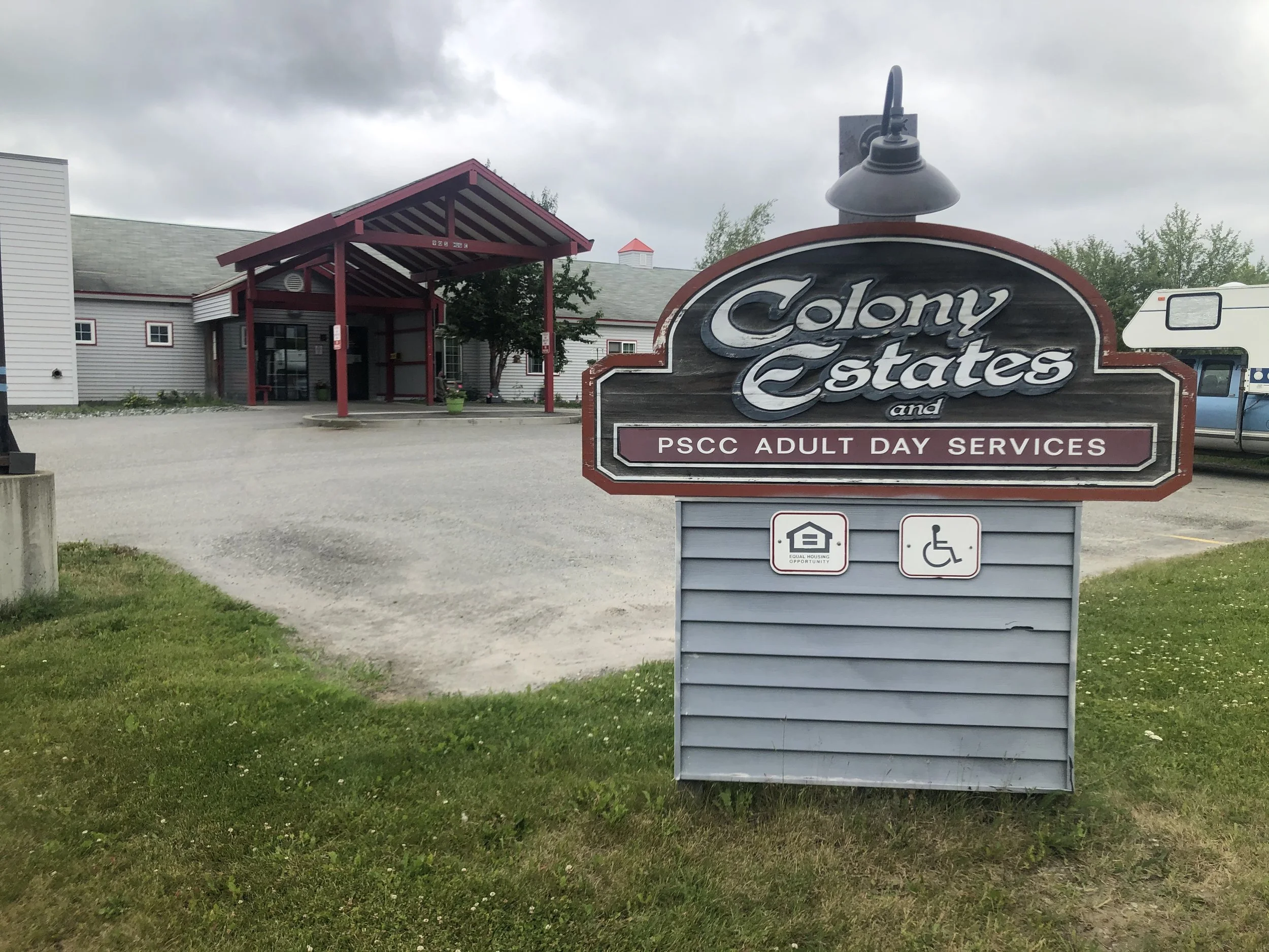 Sign for Colony Estates and PSCC Adult Day Services with icons for accessible parking and housing, in front of a building with a red entrance and a gravel parking lot.