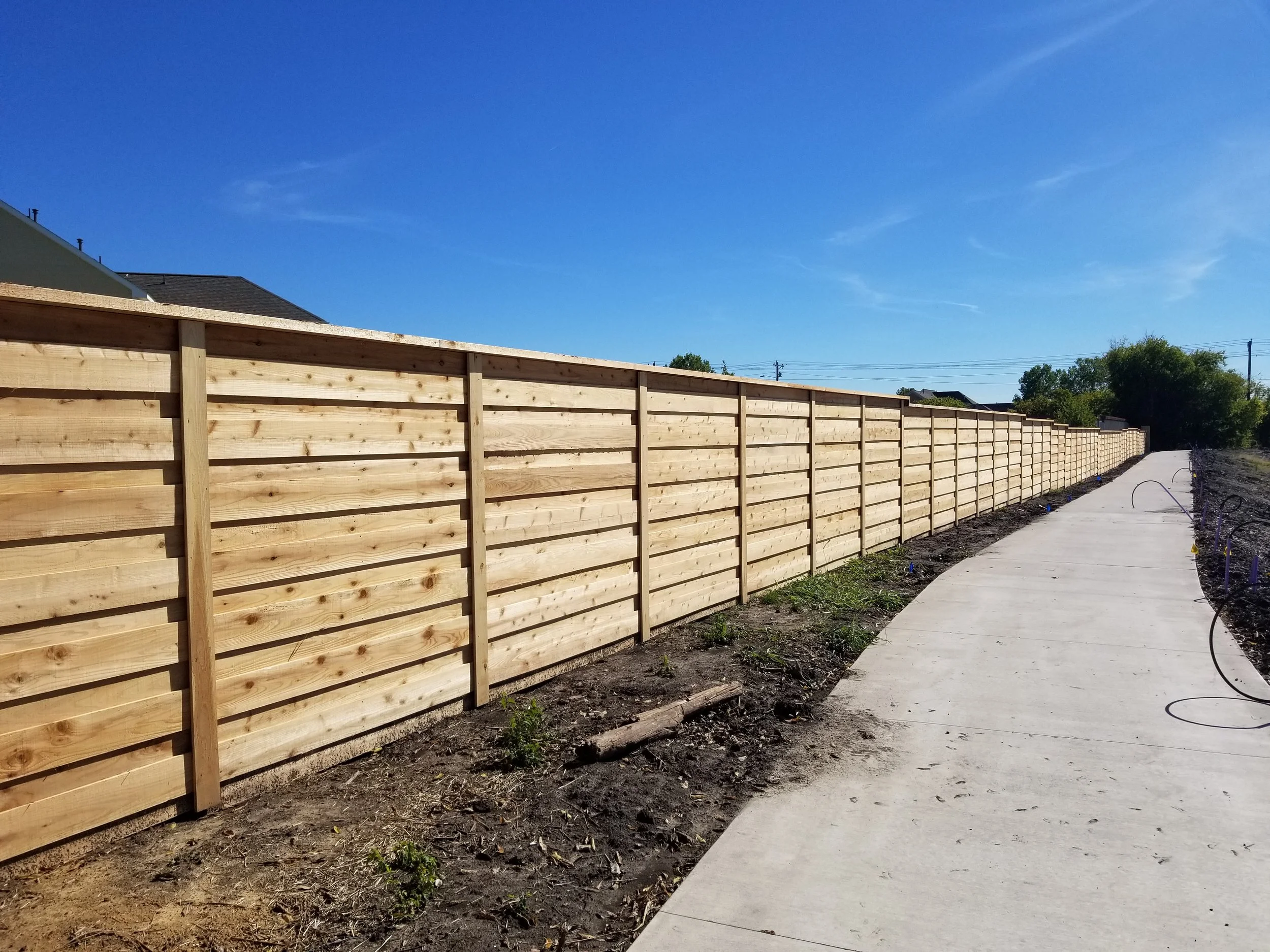 A newly installed wooden fence running alongside a concrete sidewalk on a sunny day with a clear blue sky.