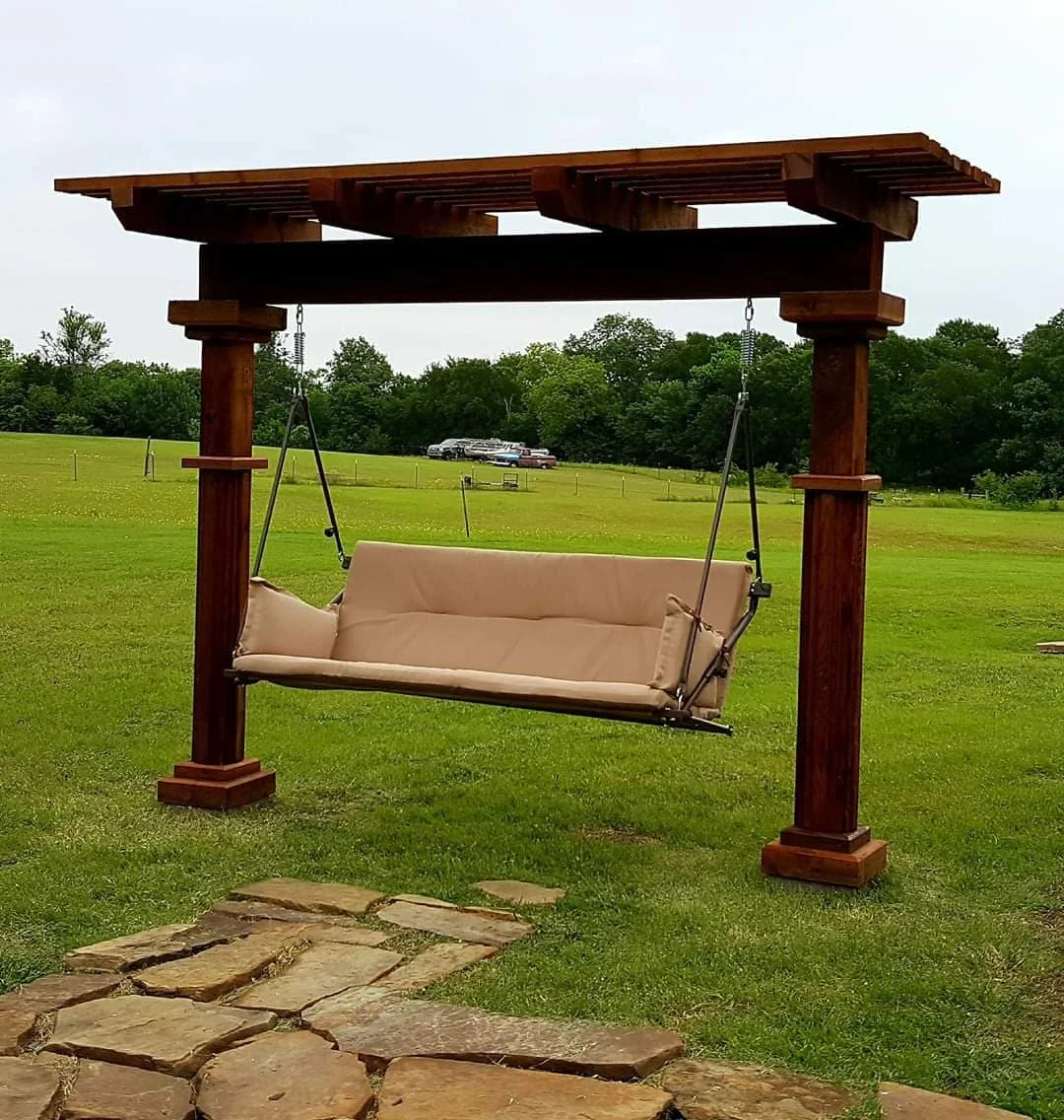 A wooden porch swing with a beige cushion hanging from a wooden frame in a grassy outdoor area, with a stone pathway leading to it and a parking lot in the distance.