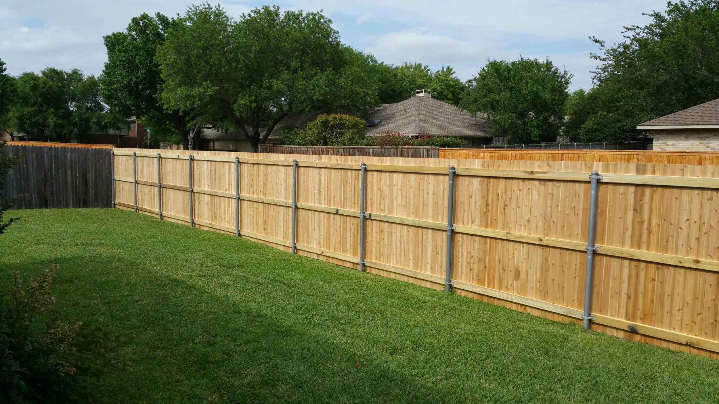 A backyard with a newly installed wooden fence, green grass, and trees in the background under a partly cloudy sky.