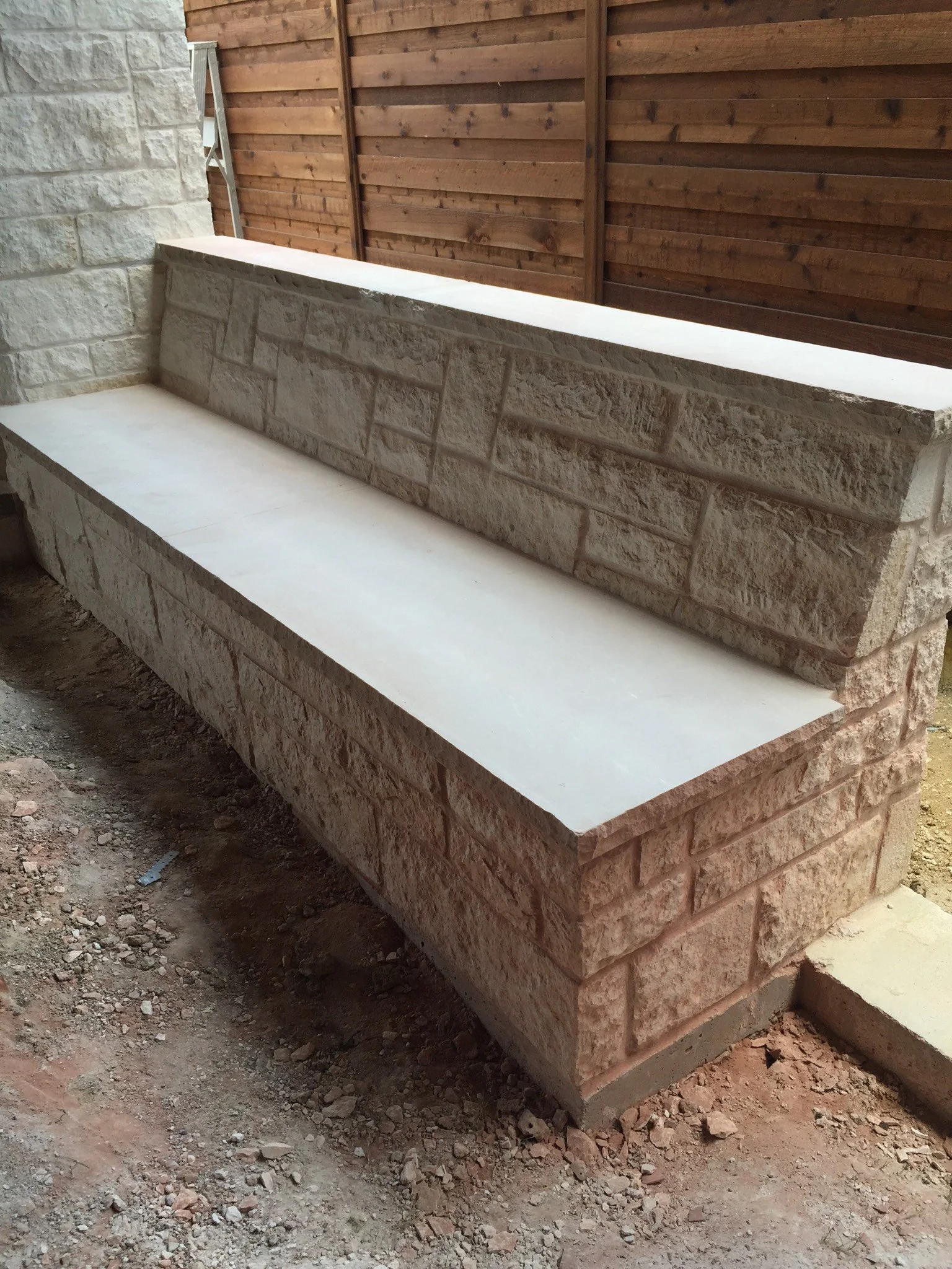 A stone bench with a white top is located outdoors near a wooden fence and stone wall.