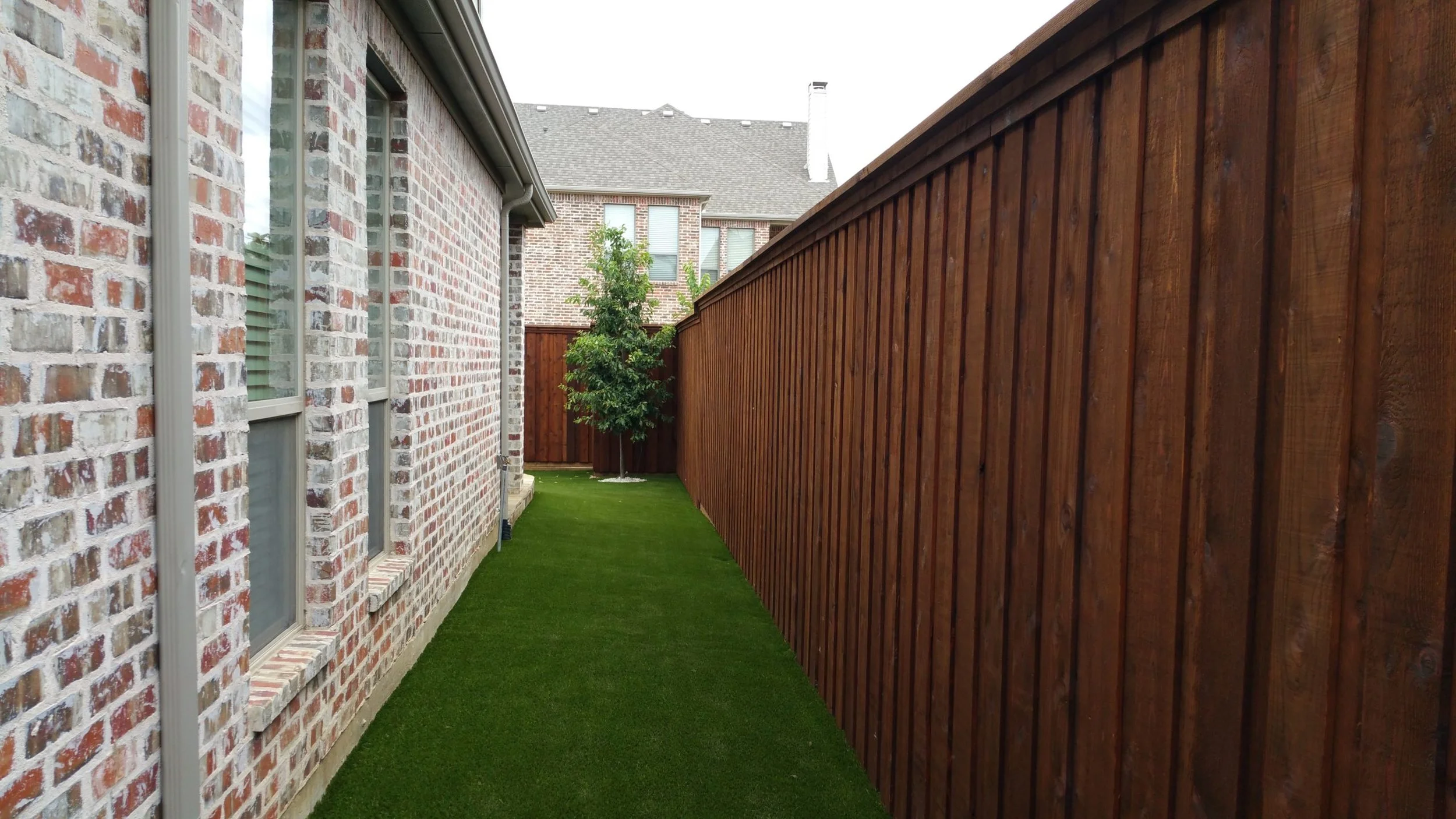 Side yard with brick house on the left, green artificial grass, and a tall wooden fence on the right. At the end of the yard, there is a small tree and taller houses in the background.