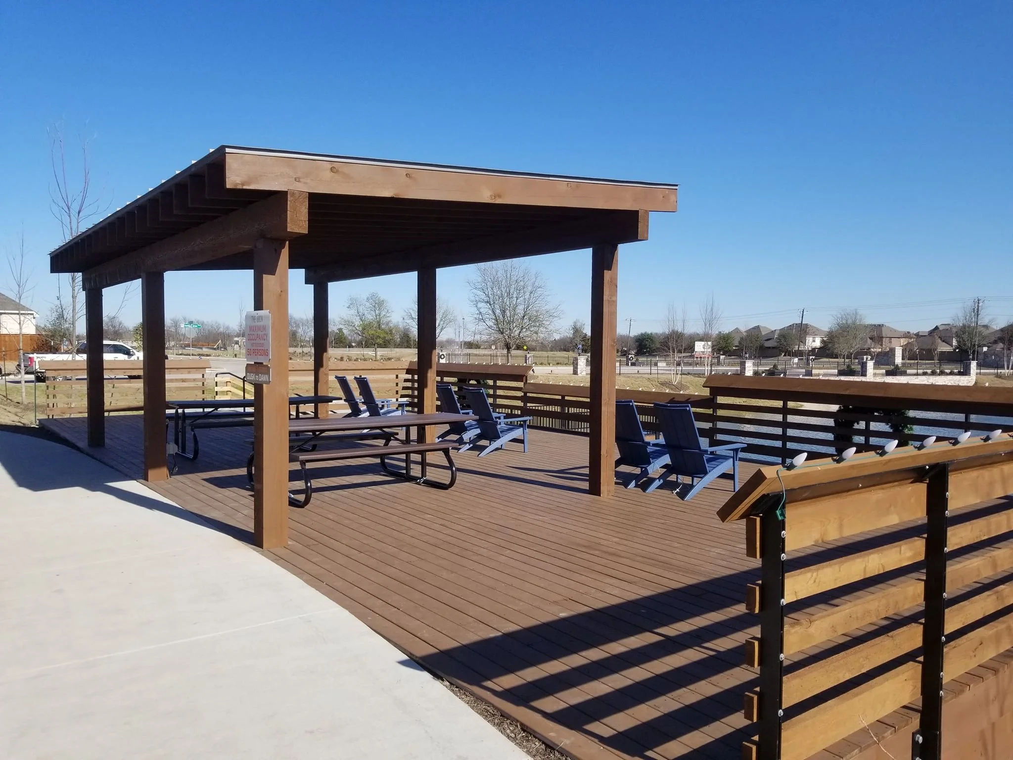 An outdoor wooden pavilion with a roof, picnic tables, and lounge chairs on a wooden deck, with a clear blue sky and some trees and buildings in the background.