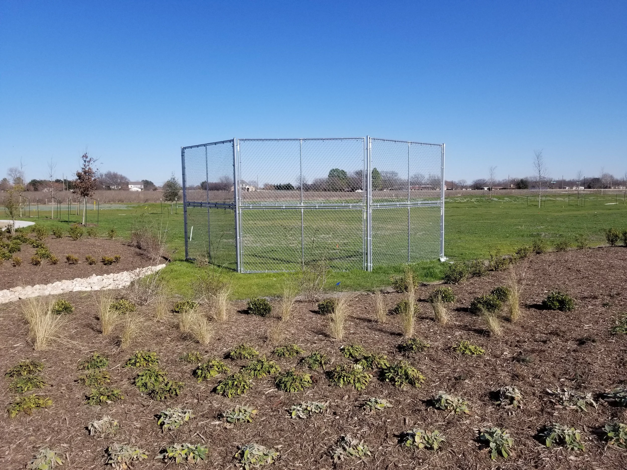 Empty chain-link fenced baseball or sports field against a clear blue sky, surrounded by grass and planted shrubs.