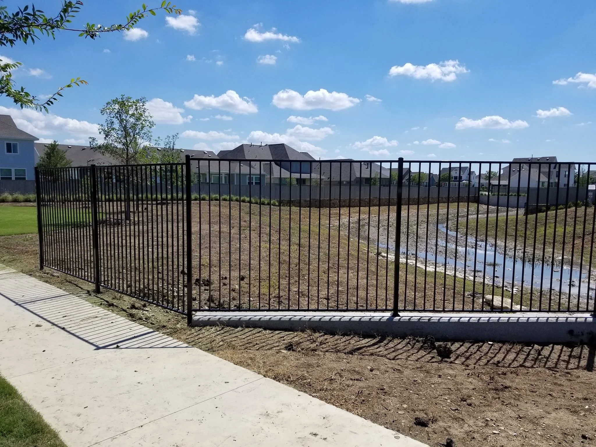 Empty fenced yard with a small drainage ditch, surrounded by residential houses under a partly cloudy blue sky.