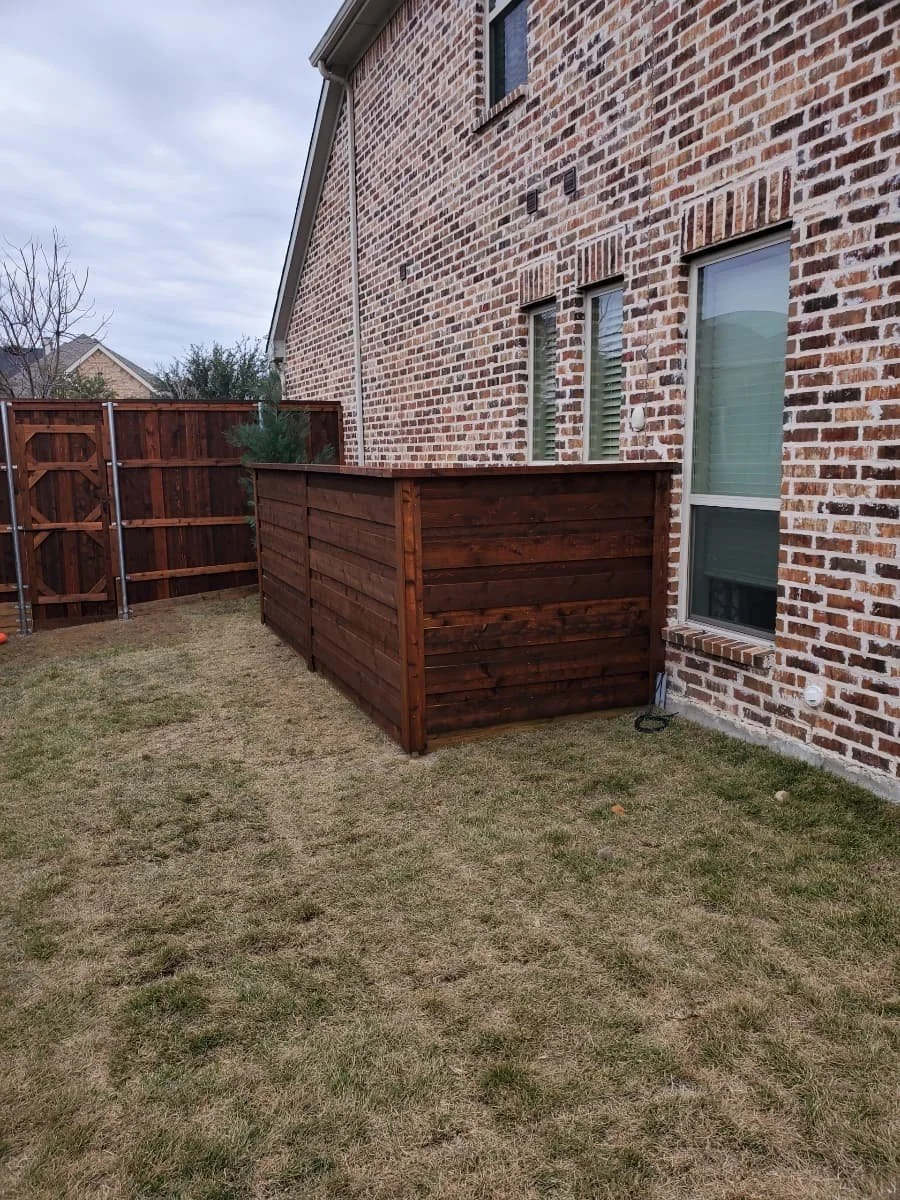 Backyard with a small wooden privacy fence next to a brick house, grass lawn, and a wooden fence in the background.