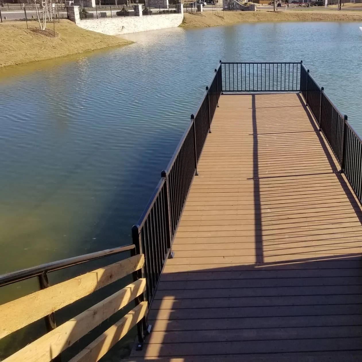 Wooden pier extending over a body of water with a black metal railing, casting shadows on the pier, in a park setting.