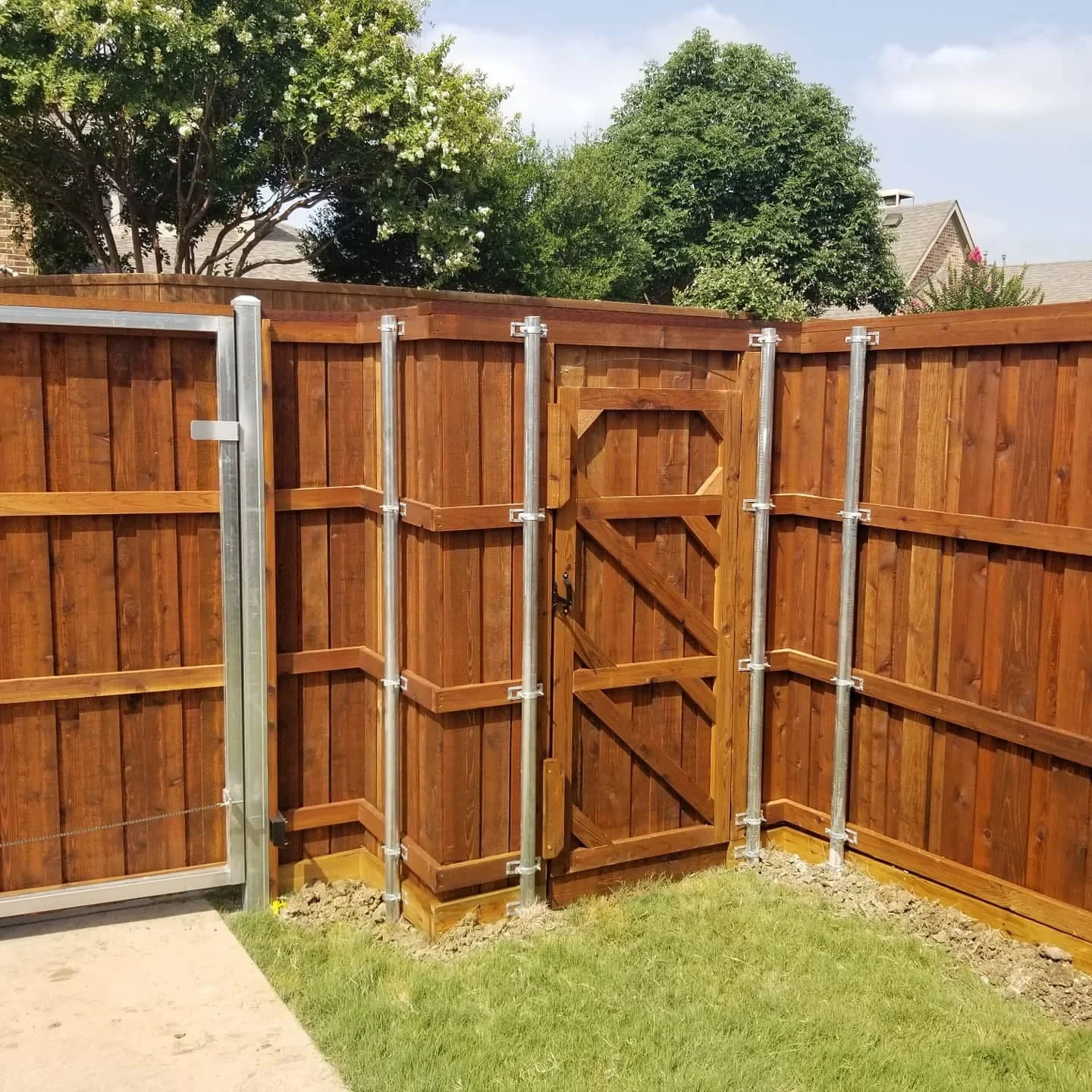 A wooden privacy fence with steel supports and a small wooden gate in a backyard with green grass and trees in the background.
