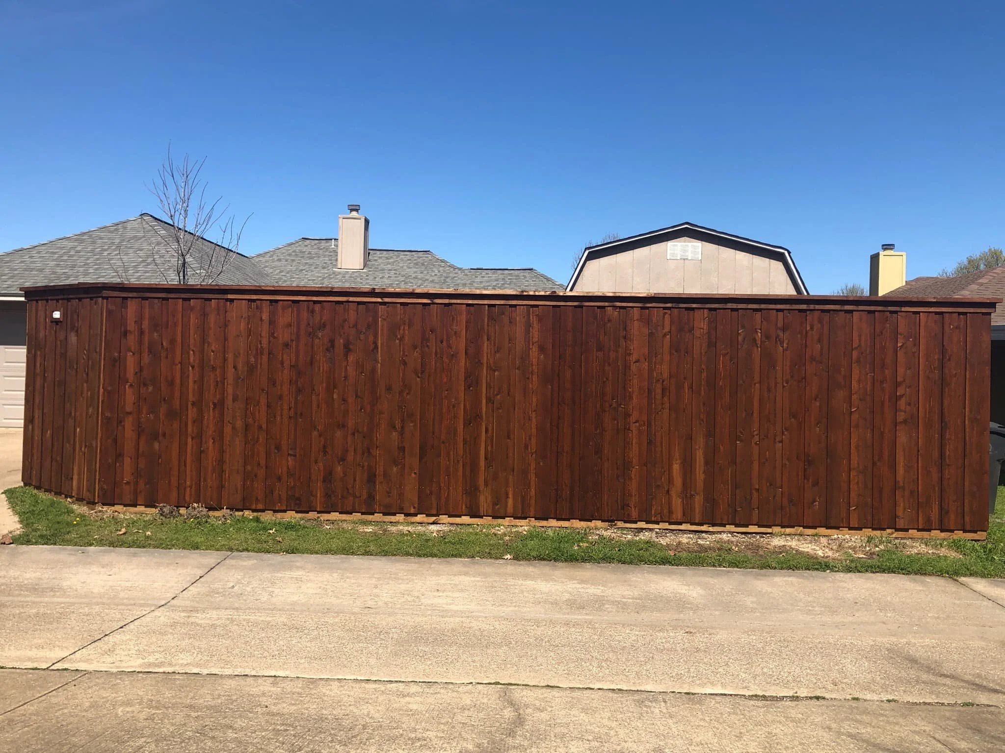 New wooden privacy fence in a residential backyard, with neighboring houses and a clear blue sky in the background.