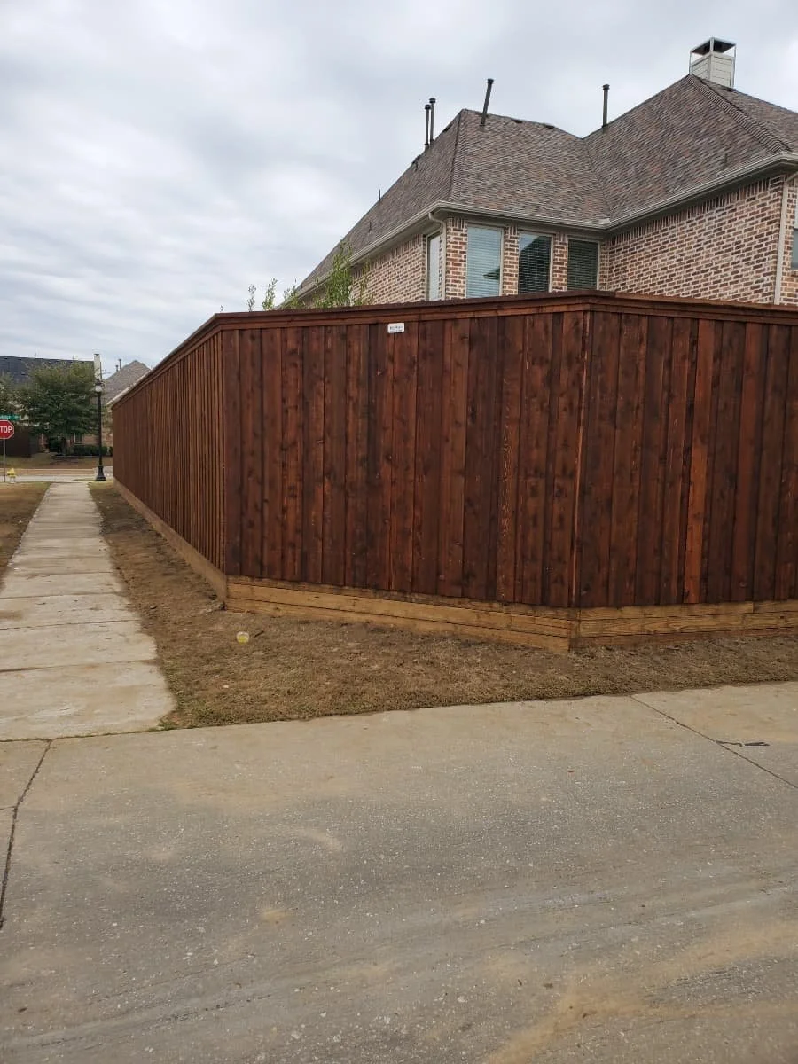 A residential backyard with a newly stained wooden fence and a brick house behind it, with a sidewalk and street in the foreground.