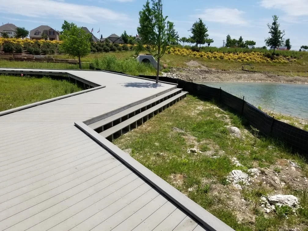 Wooden boardwalk along a grassy shoreline with trees, overlooking a body of water, with houses and flowering bushes in the background.