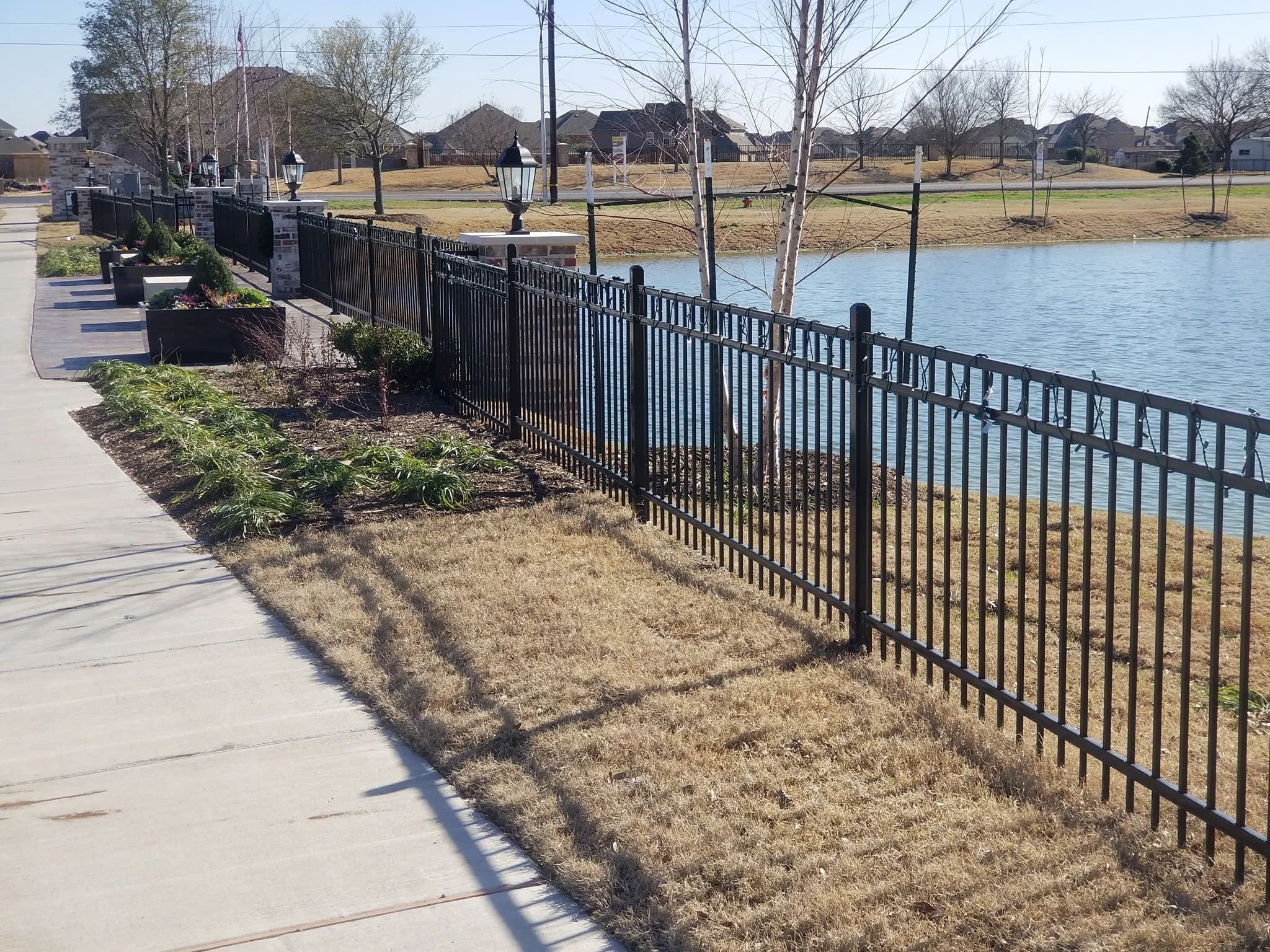 Sidewalk along a fenced pond with lawn and trees, houses in the background, and a clear sky.