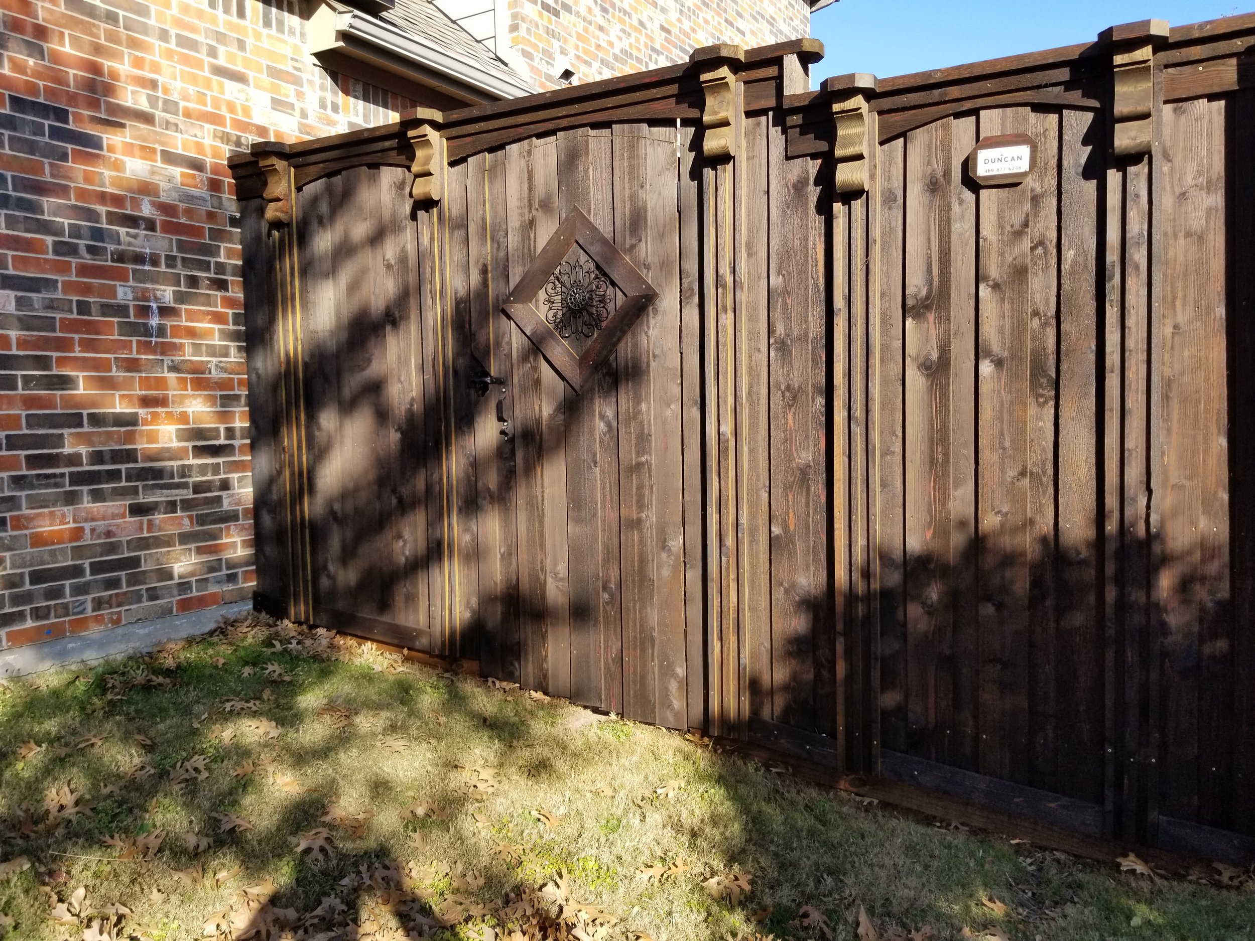 A dark wooden gate attached to a brick wall with a mailbox labeled 'Duncan' beneath a small roof. The gate has a decorative diamond-shaped window with black ironwork. There are shadows of nearby trees cast onto the gate and grass in front of it.