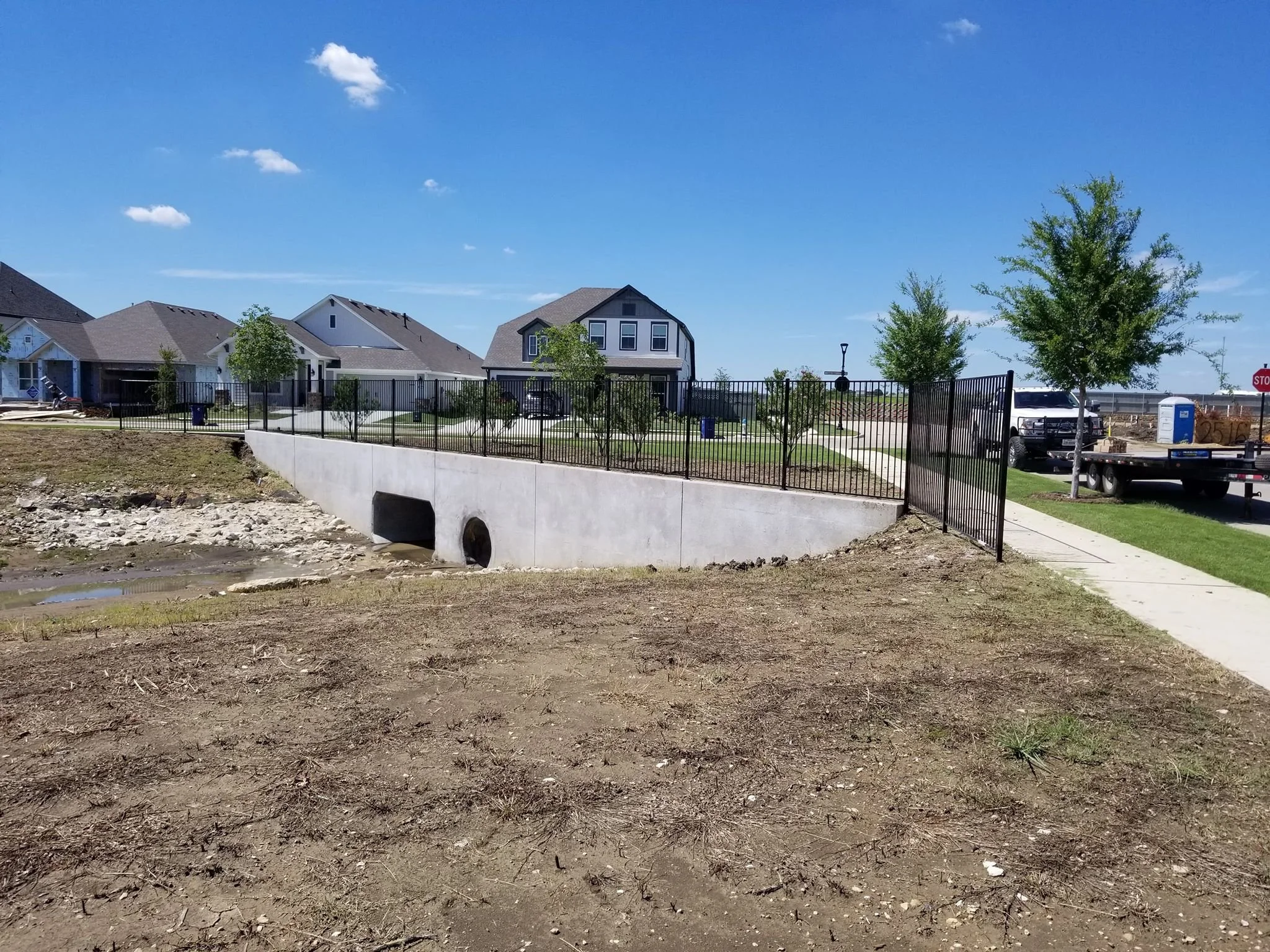 Residential neighborhood with houses, trees, and parked trucks on a sunny day. Concrete drainage structure with a black metal fence surrounding it, in the foreground.