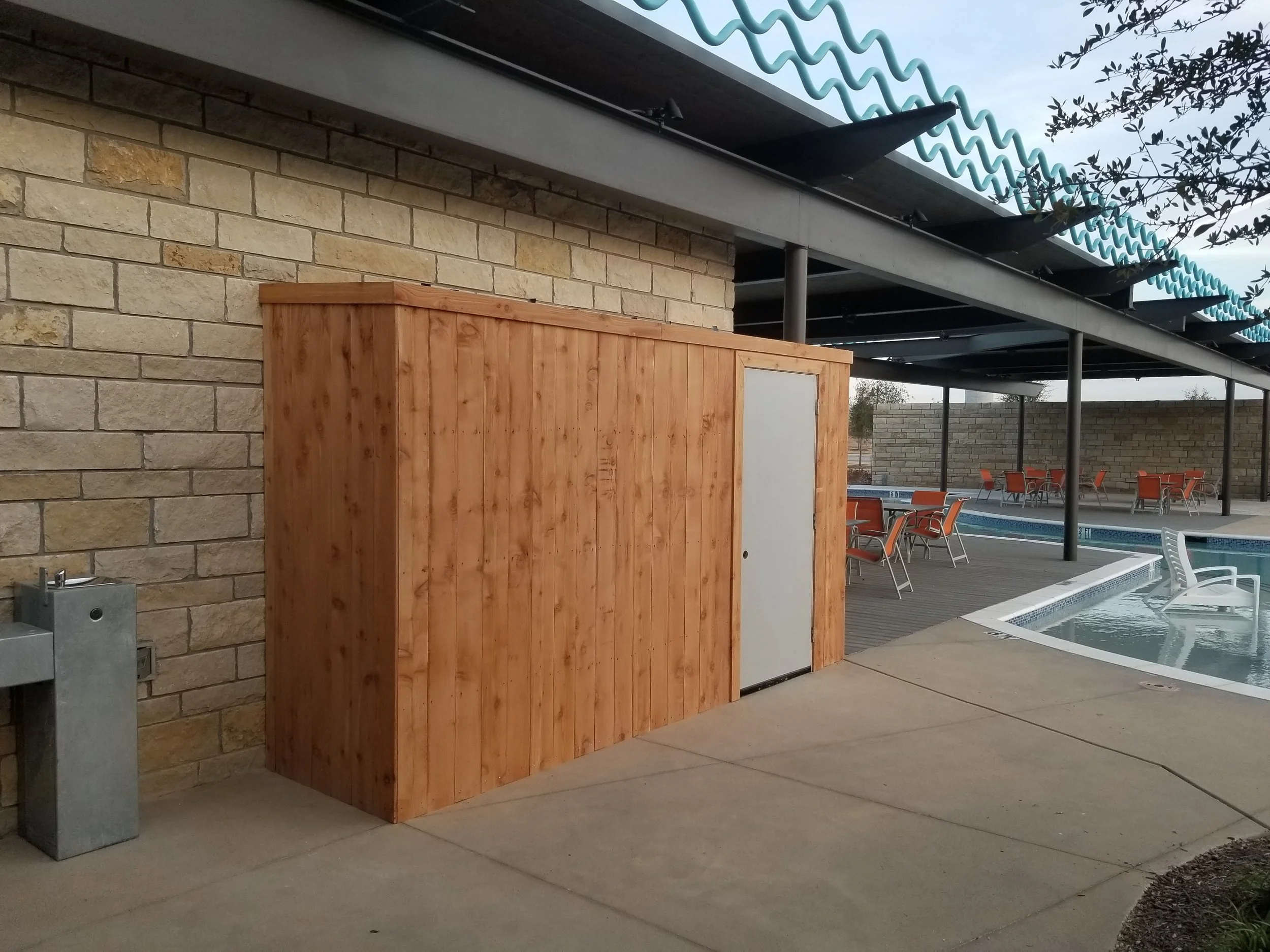 Wooden structure with a white door next to a swimming pool and outdoor seating under a covered patio.