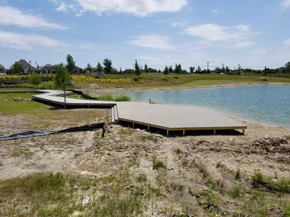 A wooden dock extending into a calm lake, with grassy and sandy shore in the foreground, and houses and trees in the background under a partly cloudy sky.