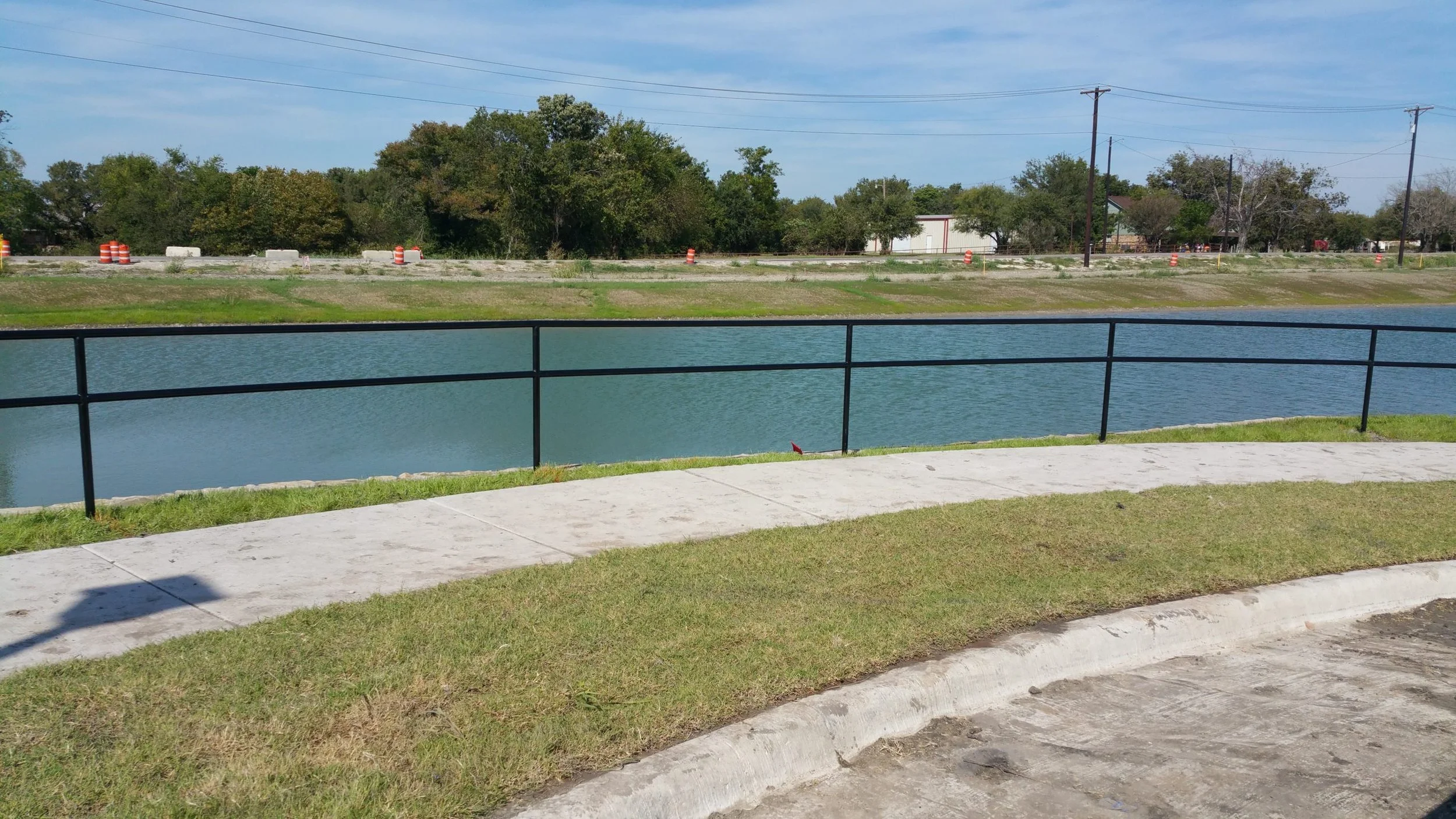 A sidewalk runs alongside a grassy area with a black metal fence overlooking a water canal, with trees and a road with orange construction barrels in the background under a blue sky with some clouds.