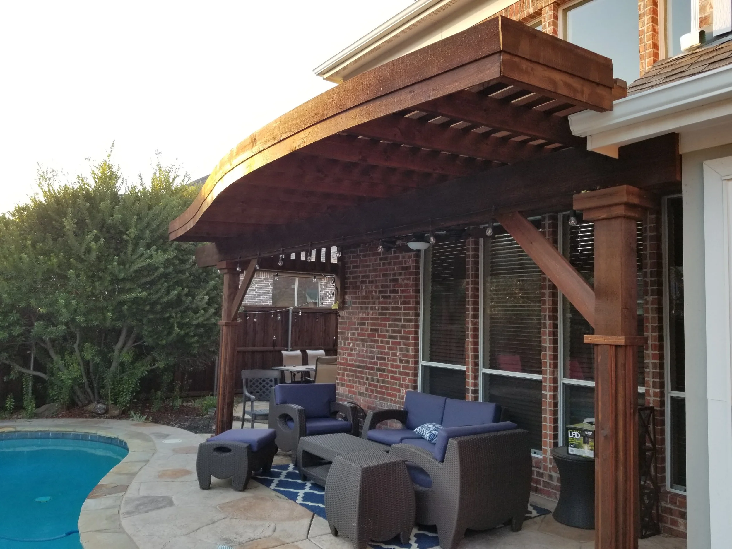 Backyard patio with a wooden pergola, outdoor seating including a love seat, chairs, and ottomans, a patterned outdoor rug, swimming pool, and a garden with trees and bushes.