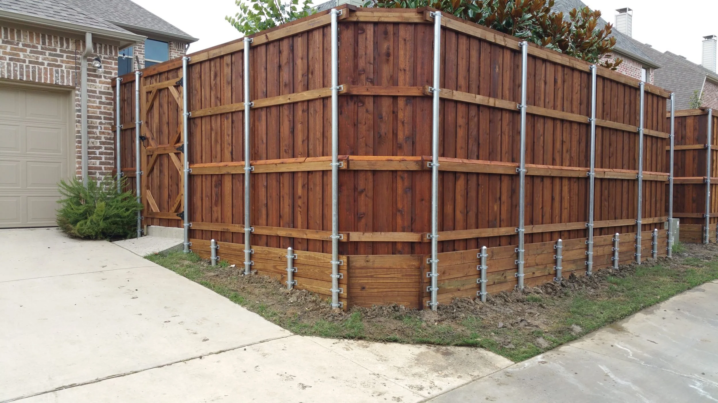 Newly installed wooden privacy fence with metal support posts surrounds the backyard corner of a house, with a driveway and neighboring houses visible.