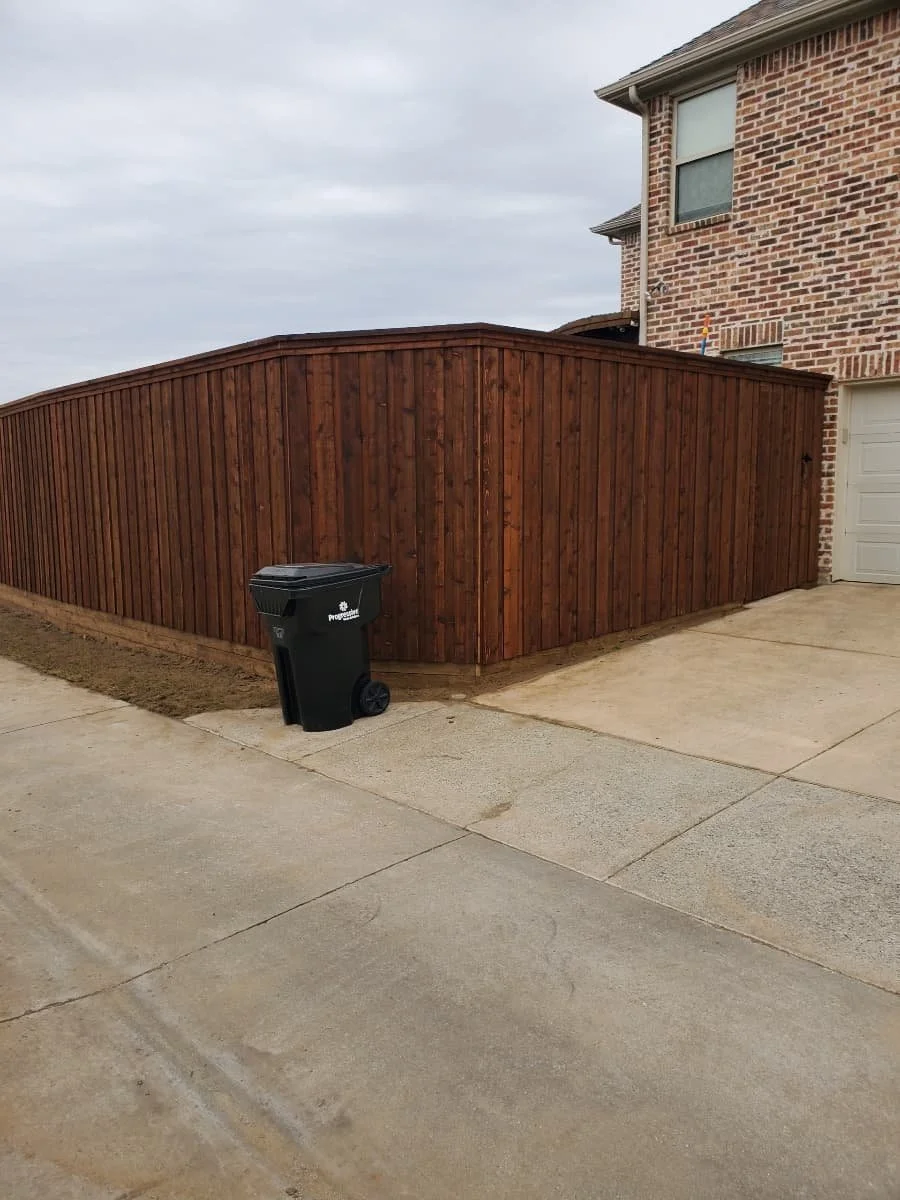 A black trash bin with wheels placed on a concrete sidewalk next to a wooden privacy fence in front of a red brick house.