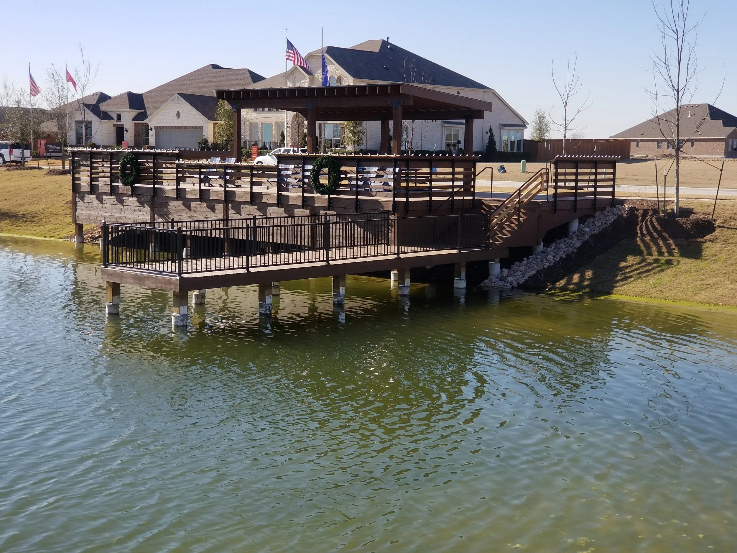 A wooden deck with black railings extends over a pond with greenish water, leading to a pavilion with holiday wreaths. Houses and leafless trees are visible in the background under a clear blue sky.
