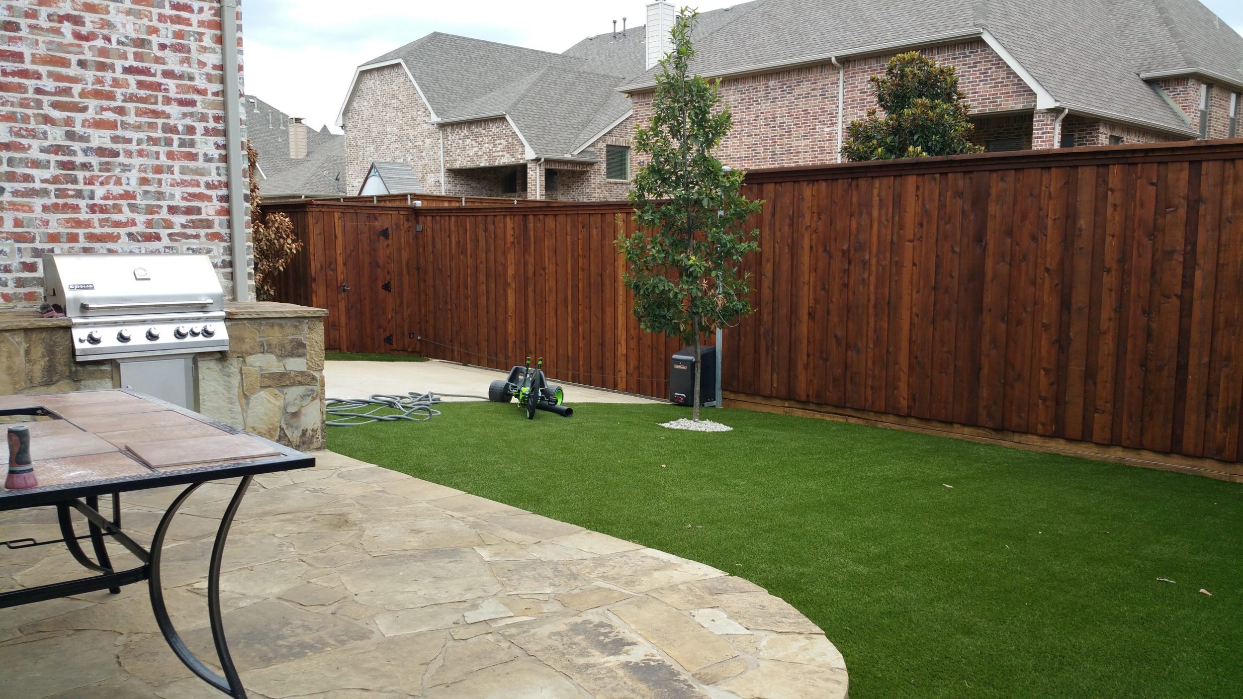 Backyard with a stone patio, a grassy area, a small tree, a wooden fence, a barbecue grill, and some gardening tools.