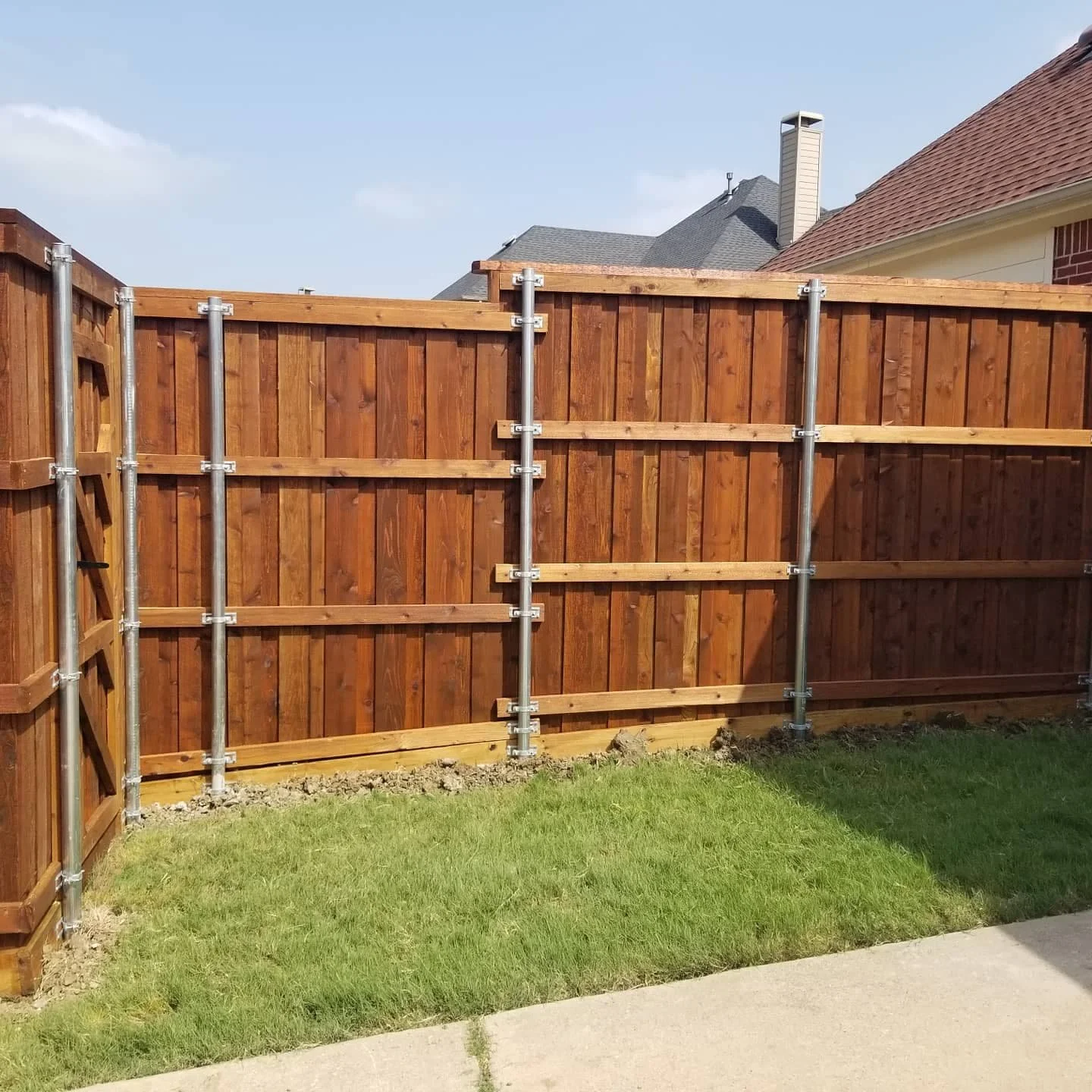 New wooden backyard fence under construction with metal posts and support beams, grassy yard, and neighboring houses visible in the background under a clear blue sky.