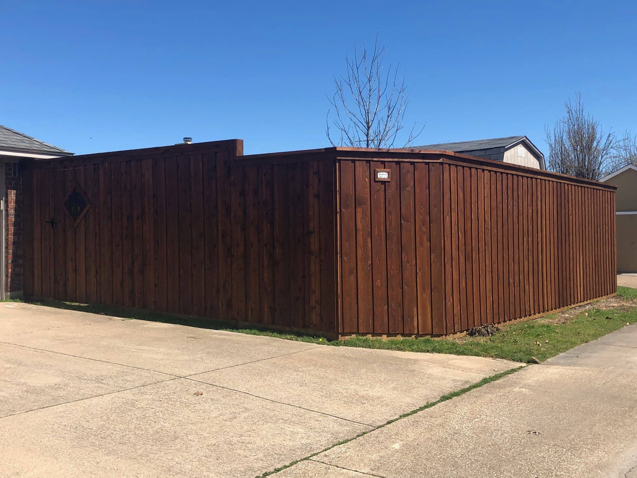 A tall wooden privacy fence surrounds a backyard, with a clear blue sky above and neighboring houses visible in the background.